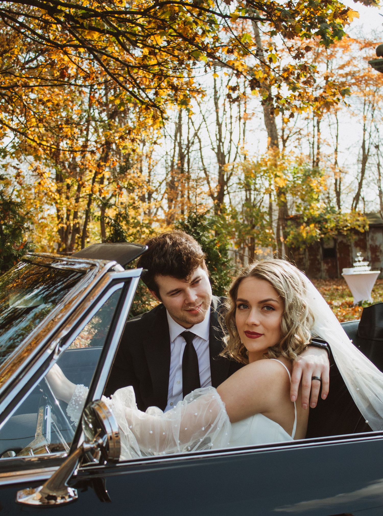 Couple in formal attire seated in vintage car during autumn.