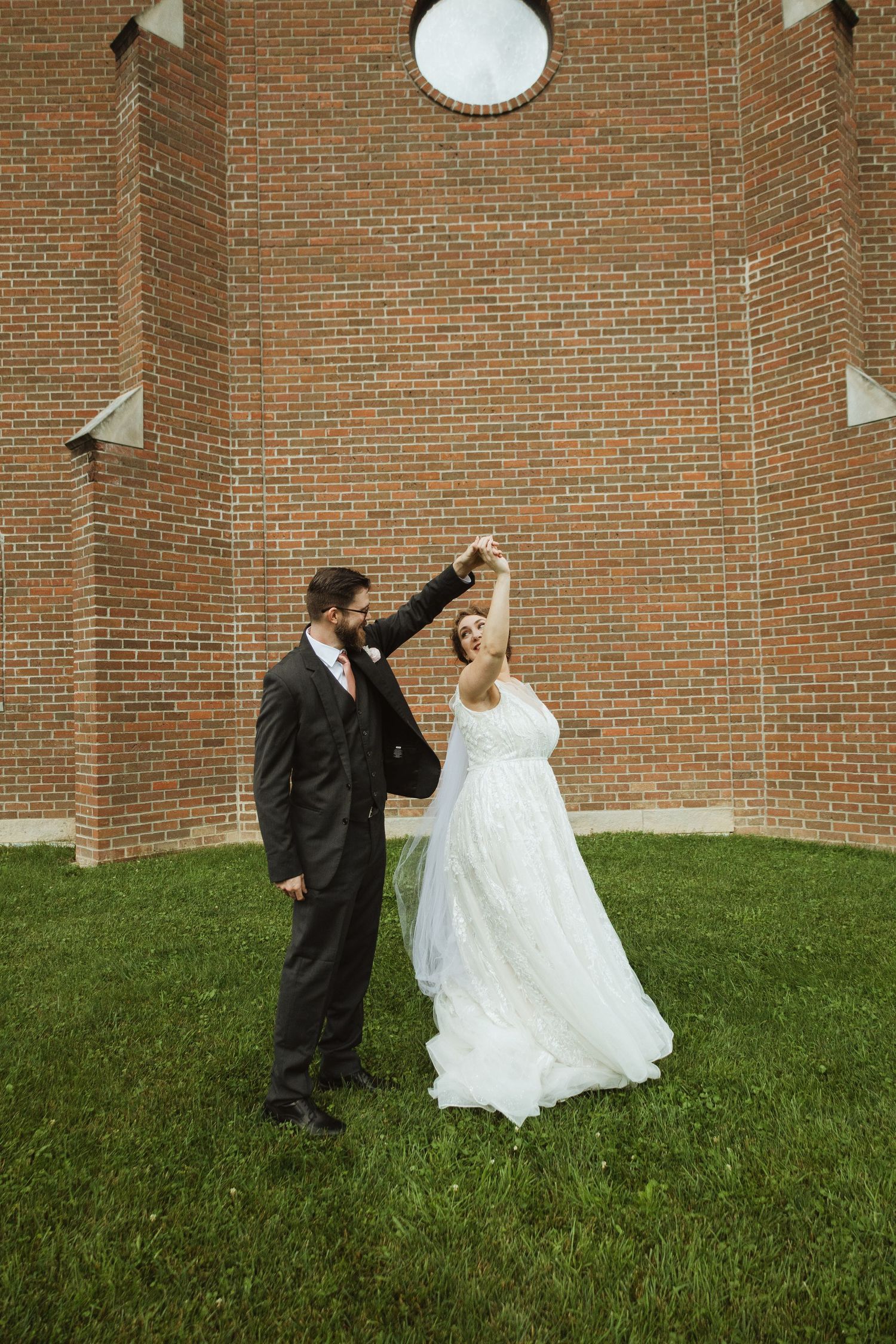 Couple dancing playfully on grass against brick wall backdrop during wedding photoshoot.