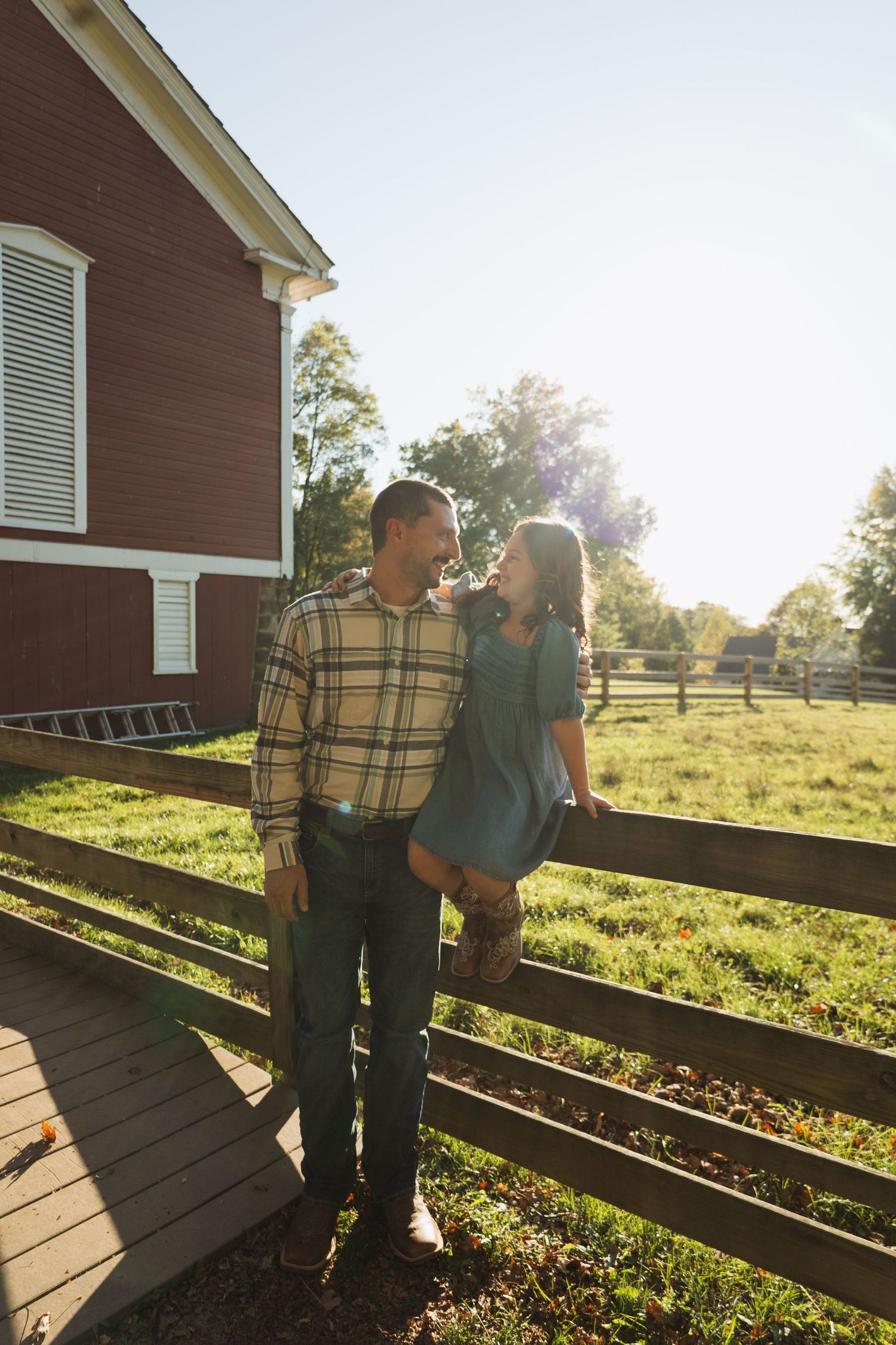 Two people stand together on a wooden deck overlooking a sunny pastoral field with wooden fencing at sunset.