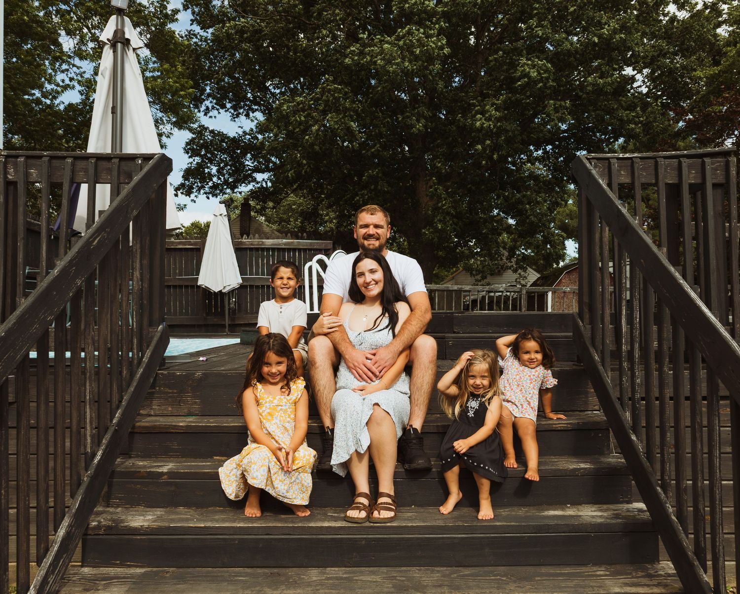 Family of six poses on wooden deck steps outdoors, with parents seated and four young daughters surrounding them.