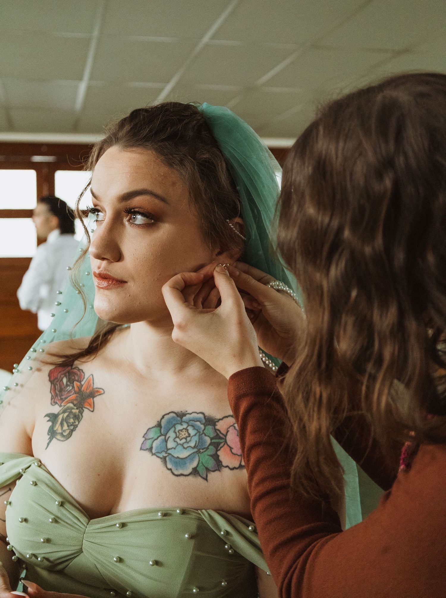 Bride in sage green dress with floral tattoos getting earring adjusted before wedding ceremony.