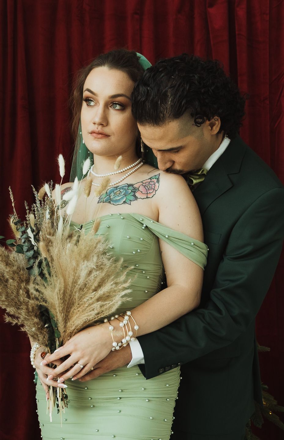 Couple in green wedding attire pose romantically against red velvet backdrop, bride holding dried pampas grass bouquet.