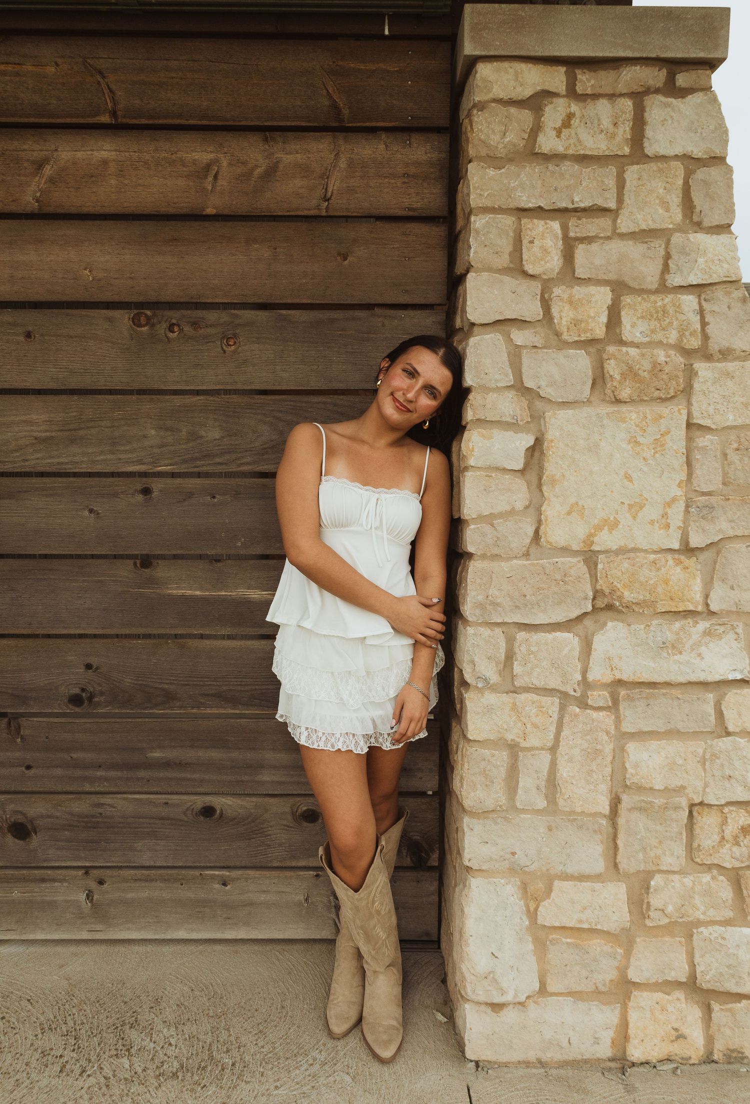 Young woman in white ruffled dress and tan cowboy boots leaning against a stone pillar near a wooden wall.