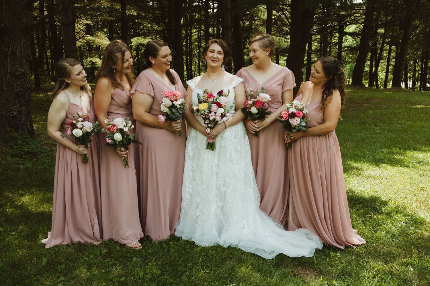 Bridal party in dusty rose dresses stands together in a scenic wooded outdoor setting.