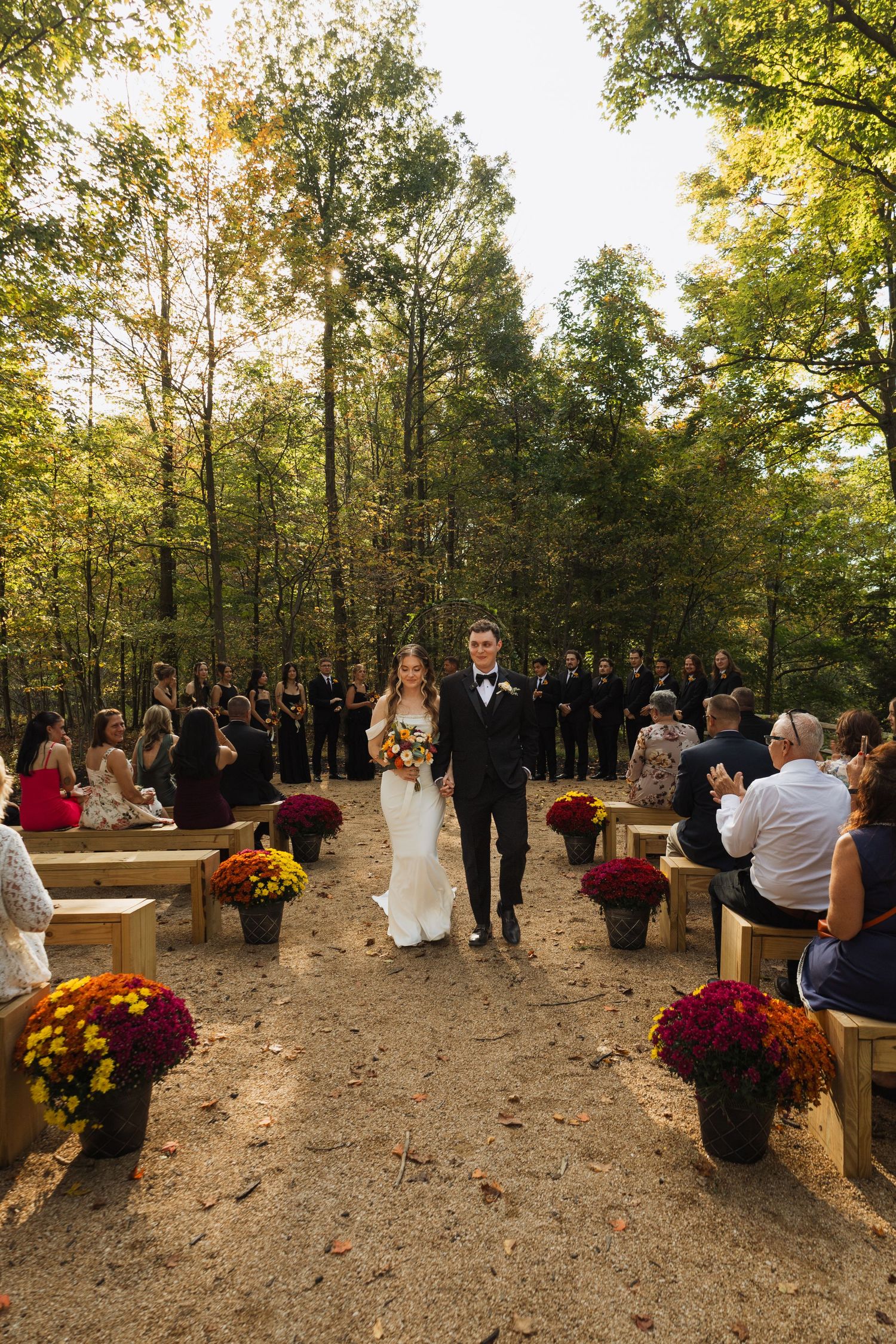 A beautiful outdoor forest wedding ceremony with guests seated on wooden benches and fall flowers lining the aisle.