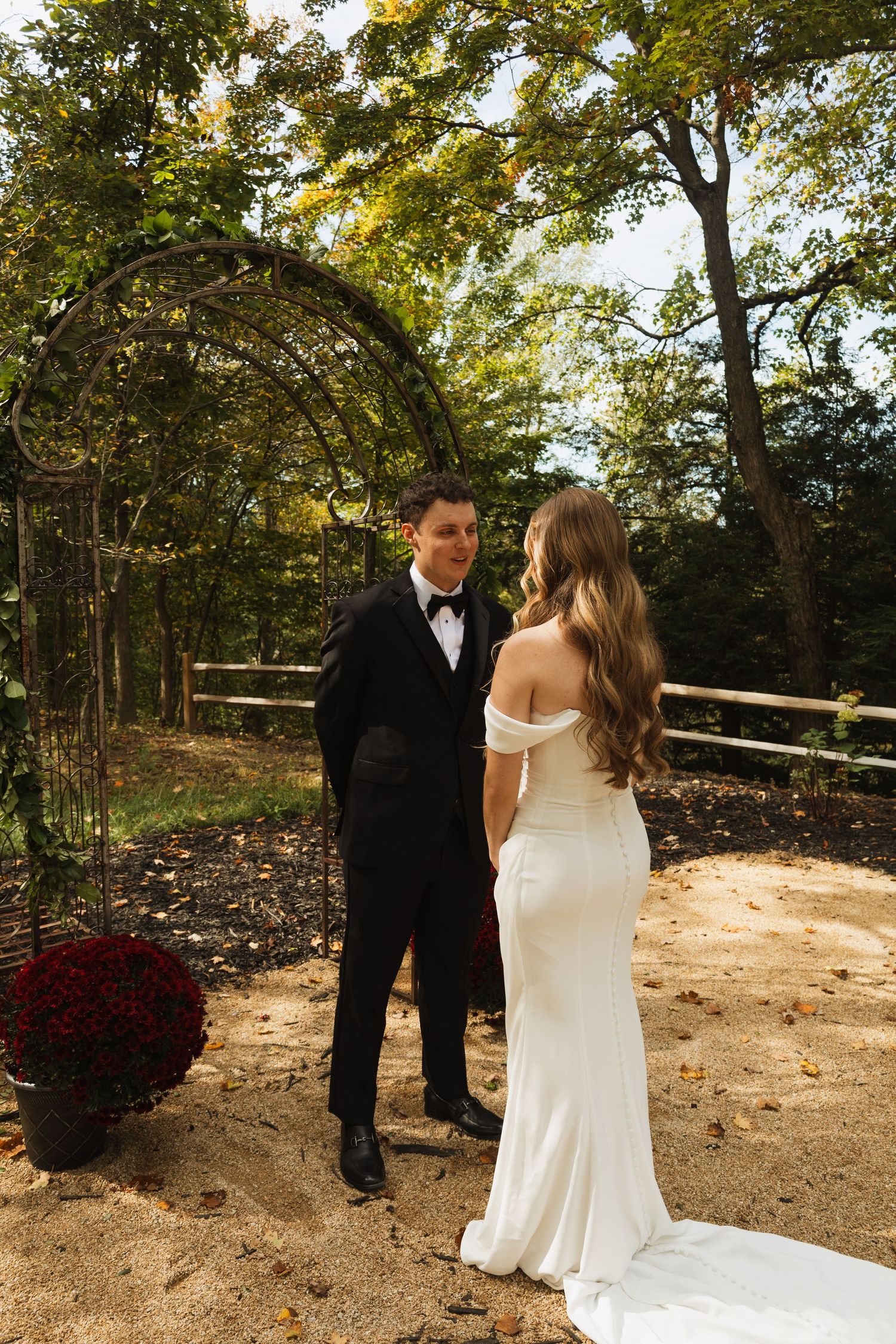 A couple in wedding attire stands together on a wooded path during golden hour.