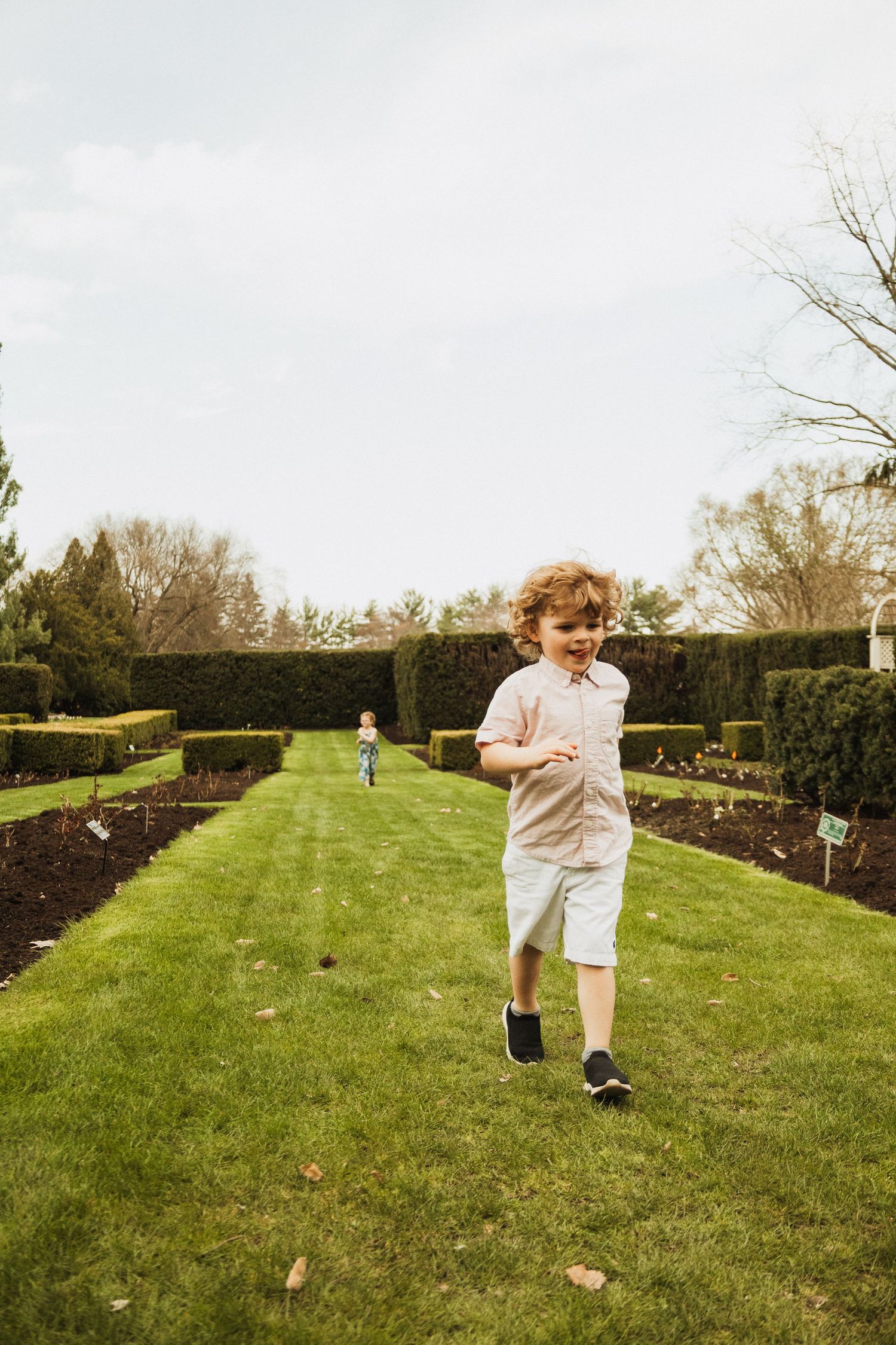 Young child running playfully on a manicured lawn path bordered by hedges in a formal garden setting.