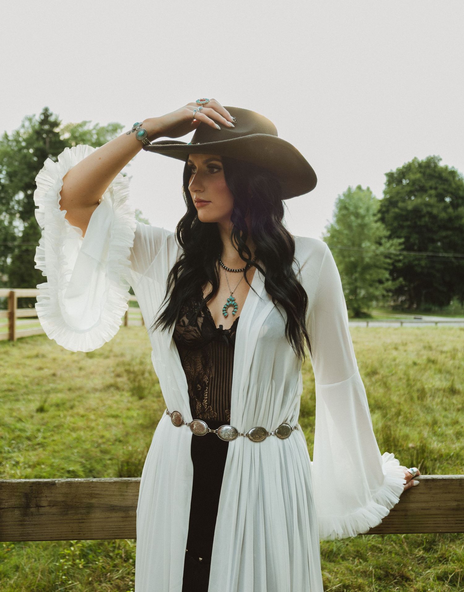 A person wearing a brown hat and flowing white dress poses gracefully in a rustic outdoor setting with green fields.