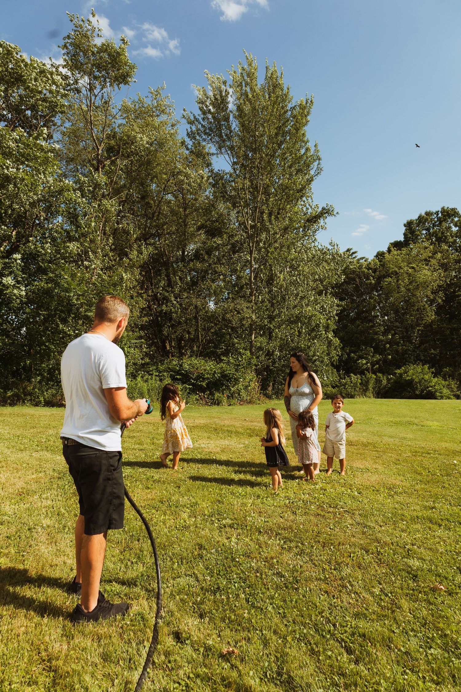 Family enjoying a sunny day outdoors on green lawn with garden hose, trees in background.