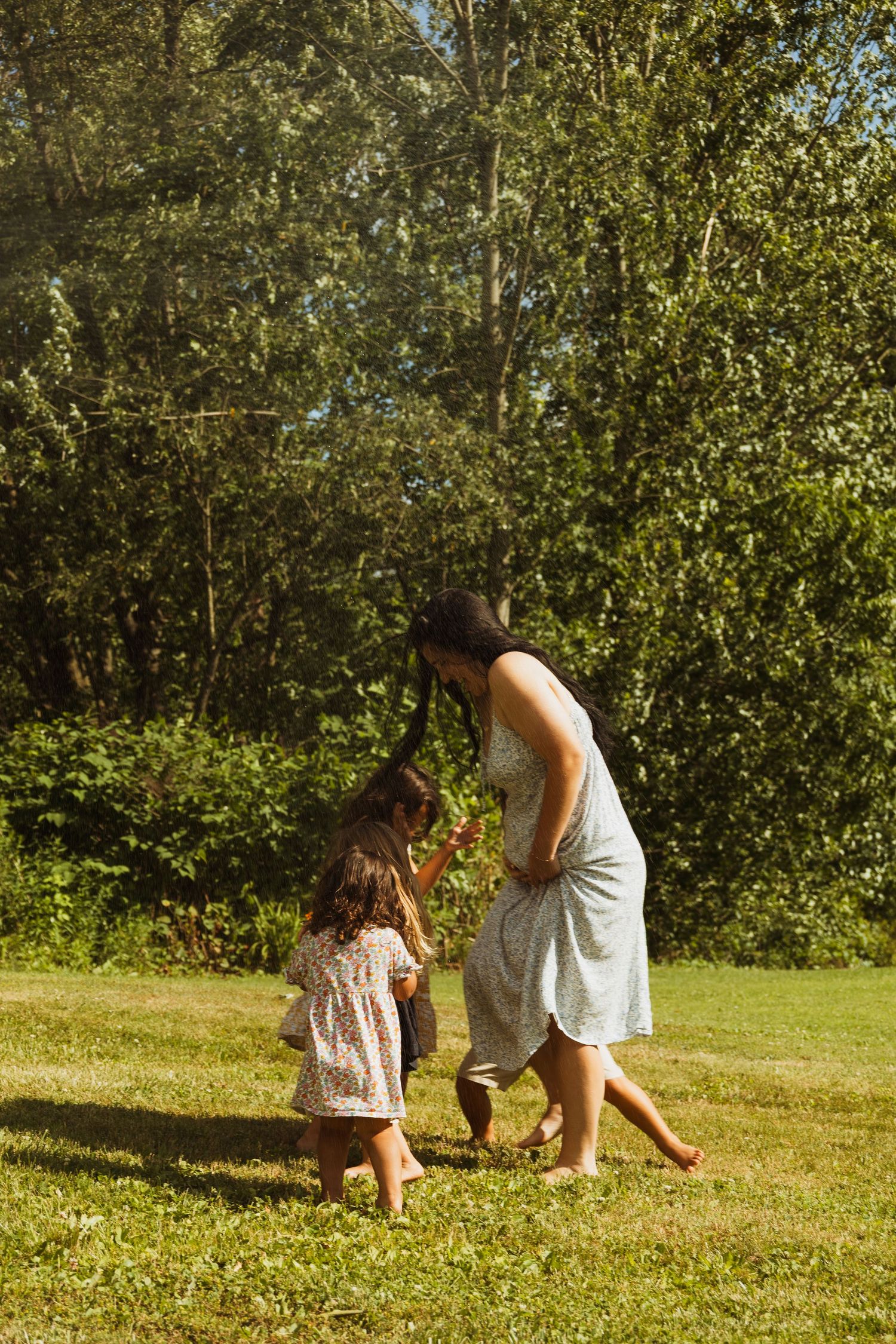 Mother and young daughter playing together on green grass in sunny backyard surrounded by lush trees.