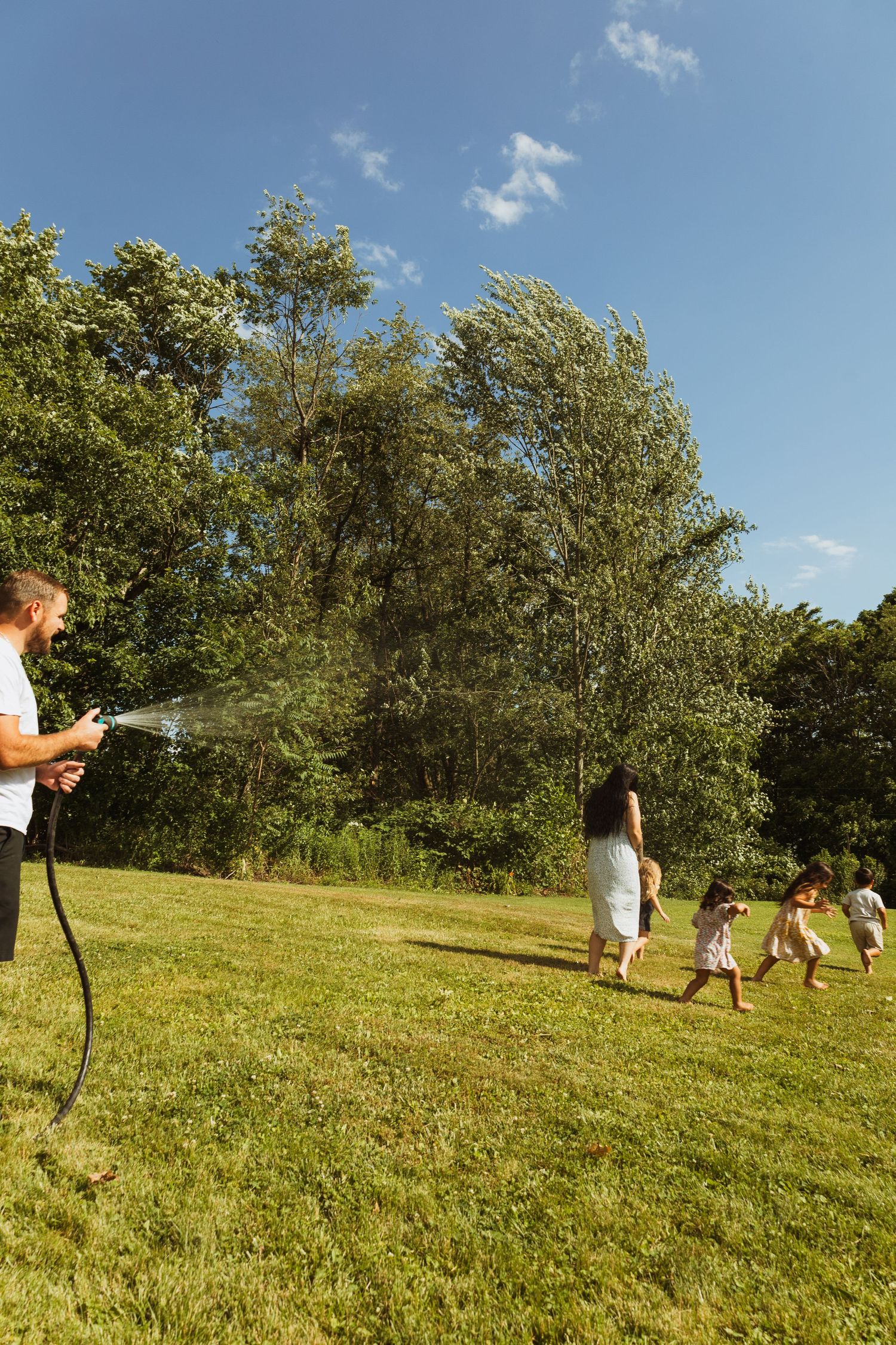 A man sprays children with a garden hose on a sunny day in a lush green park.
