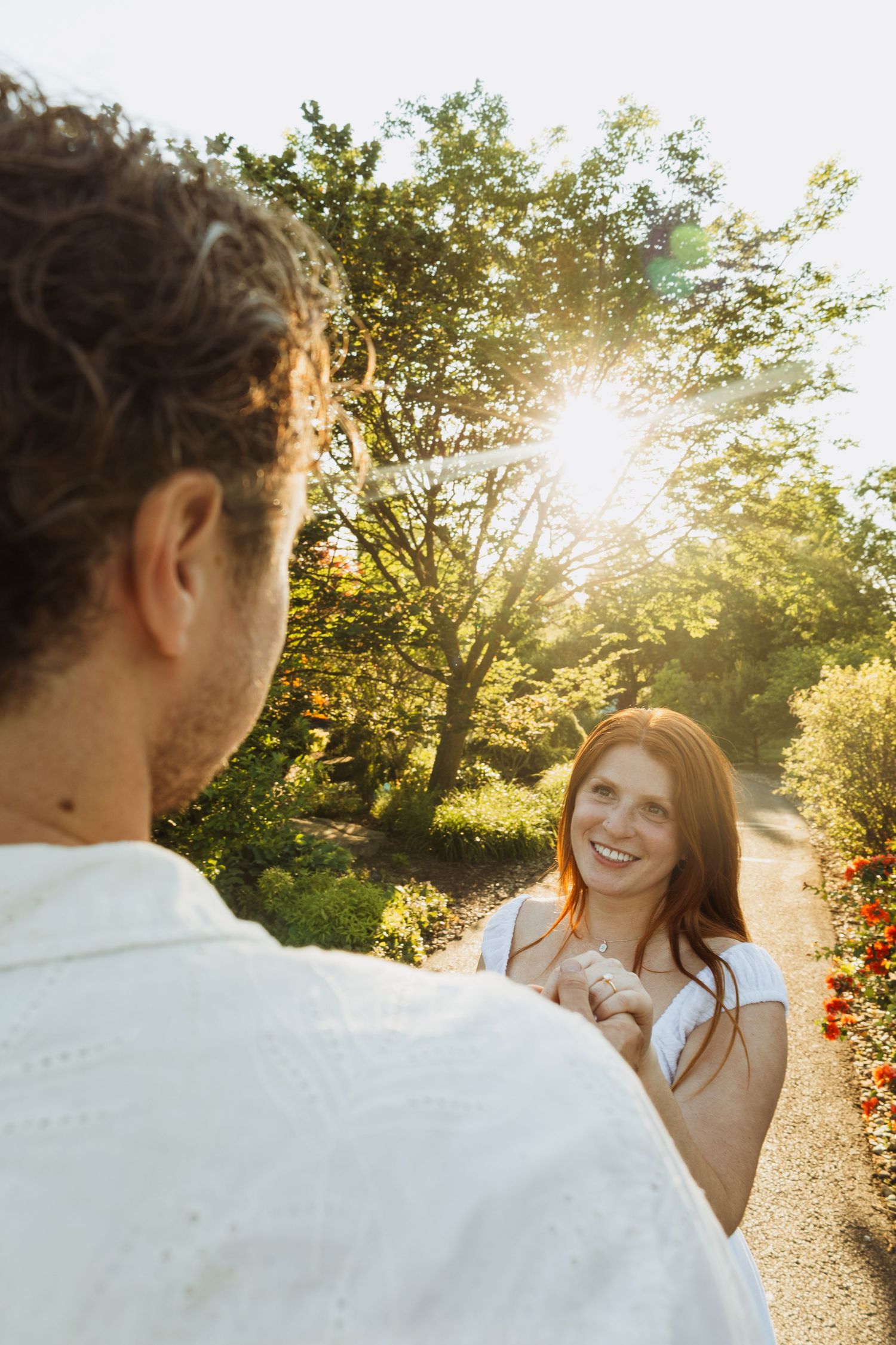 Smiling redhead woman holding hands with a man in a sunny garden park with lush green trees and flowers.