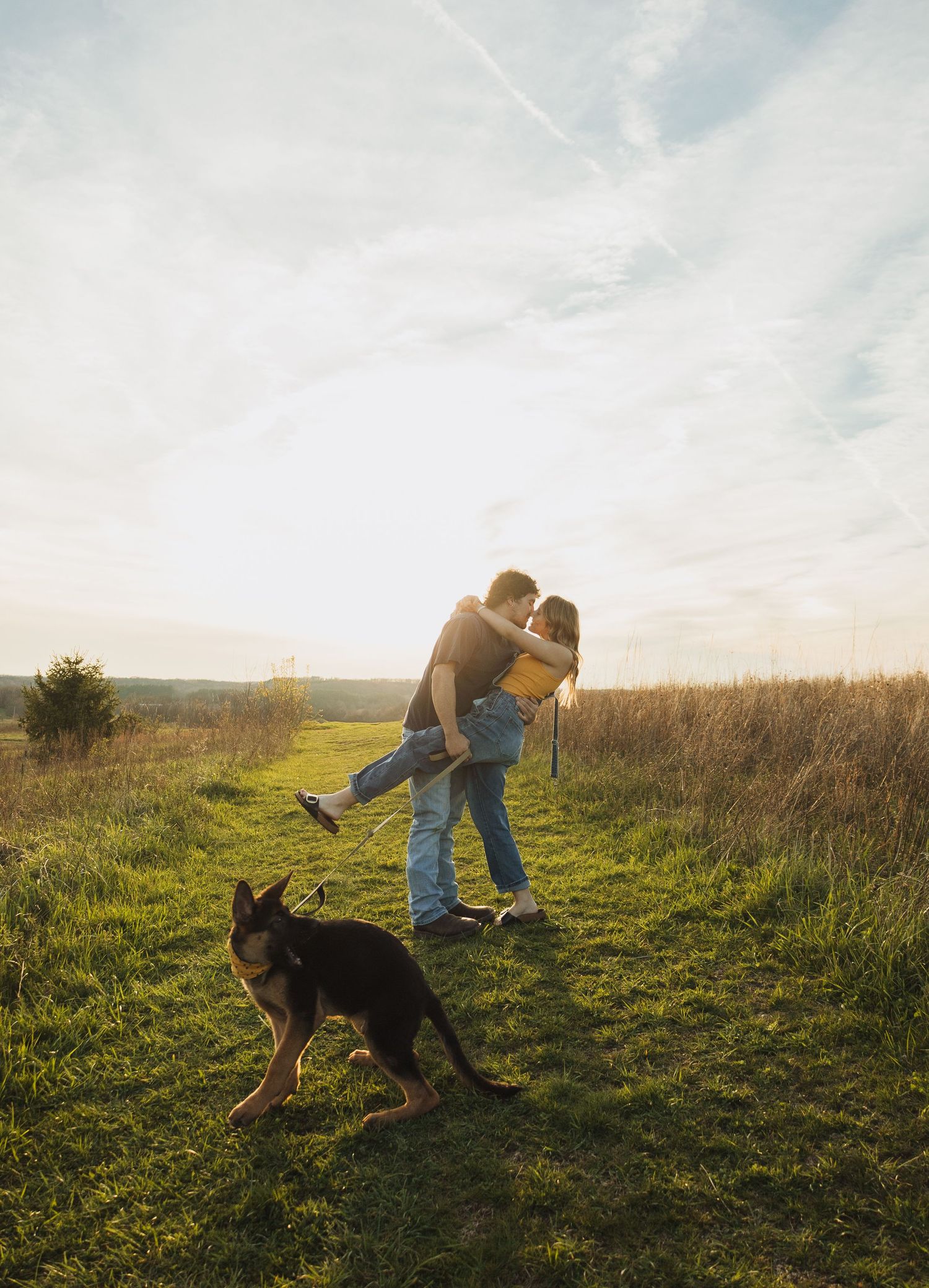 Couple embracing and kissing in a sunny green field at sunset with a German Shepherd dog in the foreground.