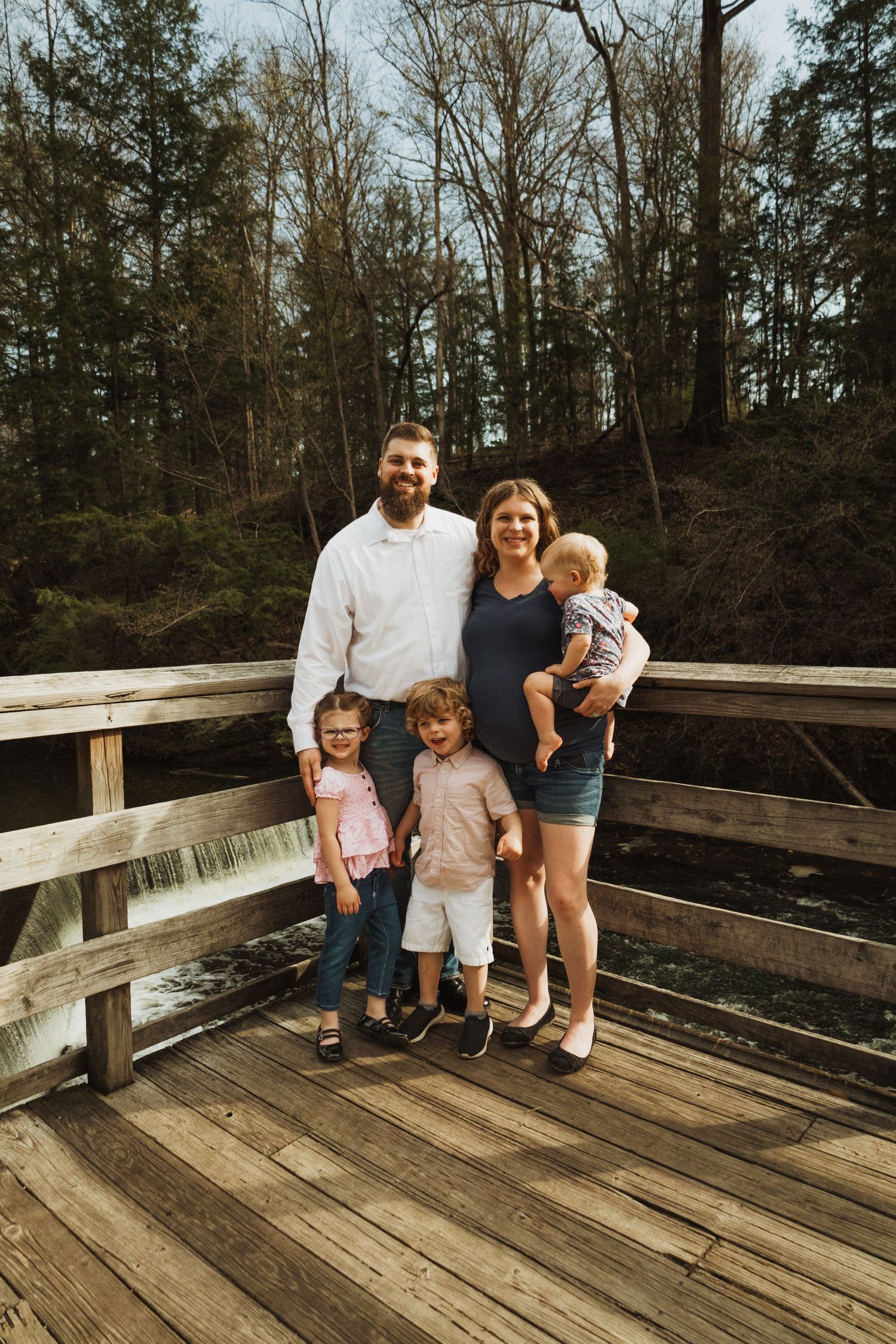 A group stands together on a wooden deck bridge surrounded by tall forest trees on a sunny day.