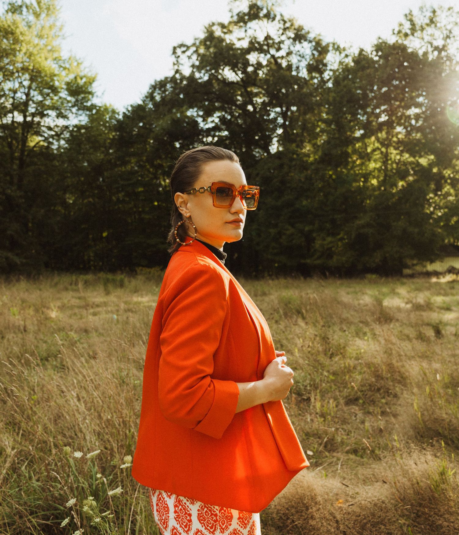 Stylish portrait showcasing orange blazer and statement sunglasses against natural outdoor backdrop.