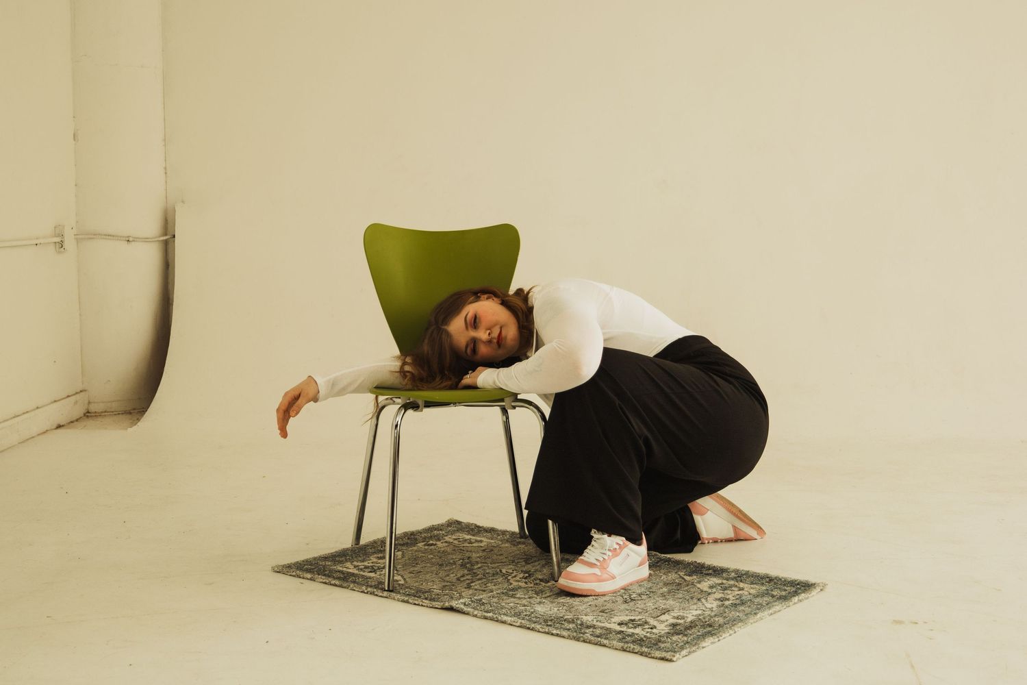 Artistic pose with person draped over green chair on patterned rug in white studio.