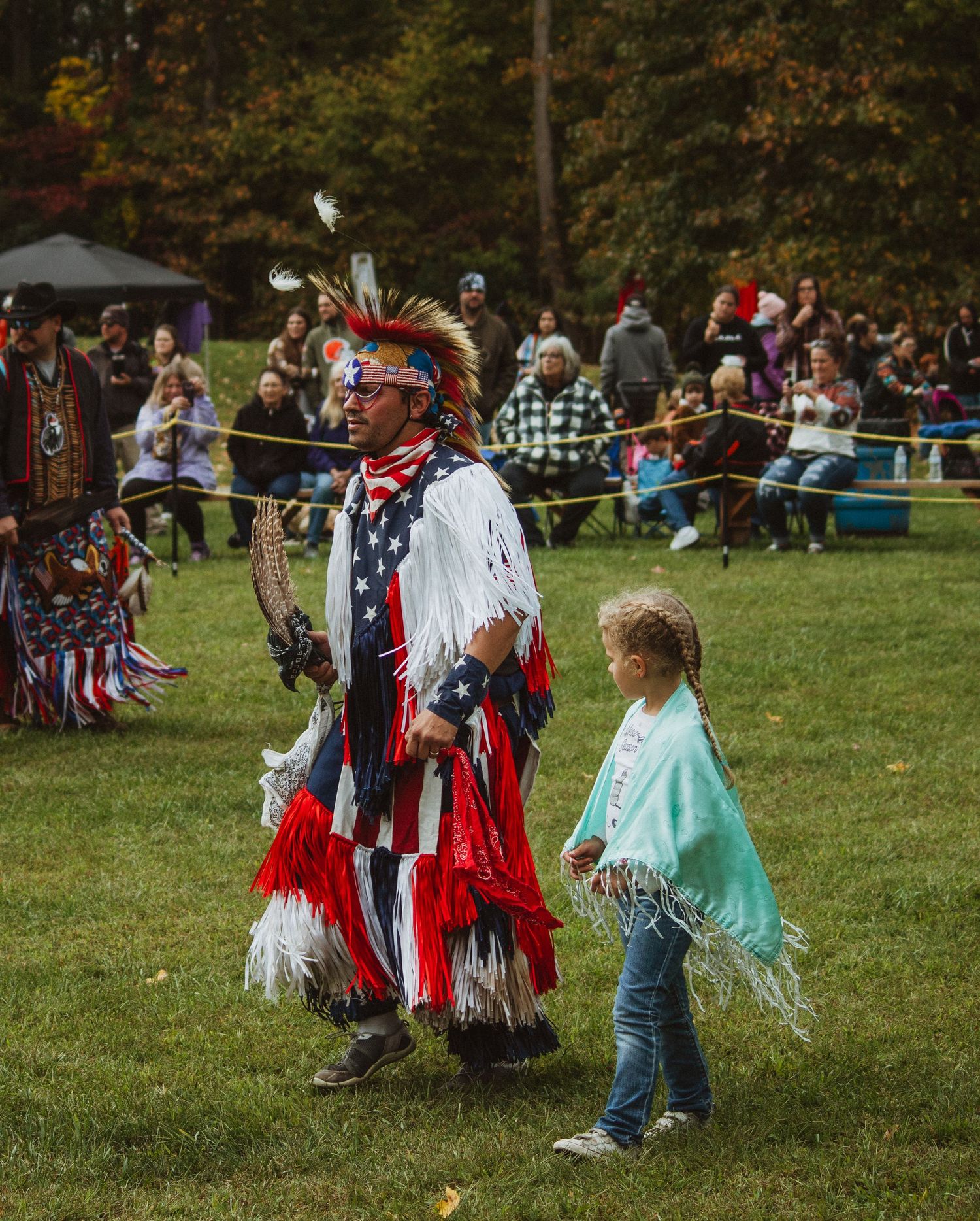 A dancer in red, white and blue regalia performs at an outdoor powwow with spectators watching.