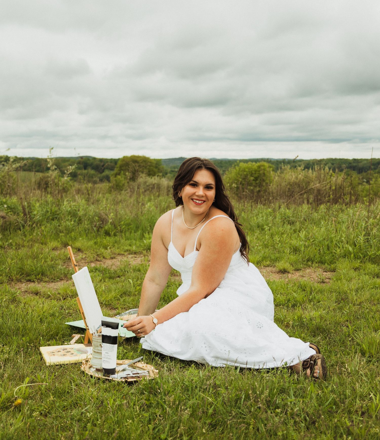 Person in white dress lying on grassy meadow with laptop and takeout food in rustic outdoor setting.