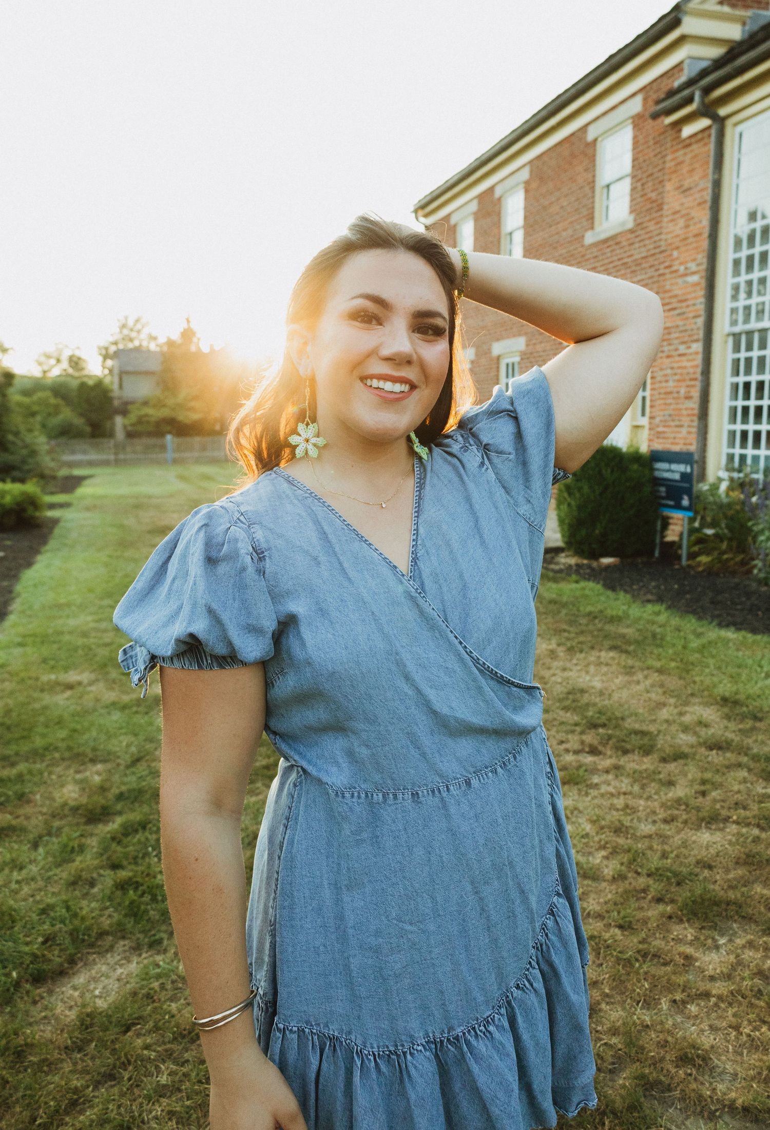 Woman wearing ruffled blue denim dress poses playfully outdoors at sunset near a brick building.