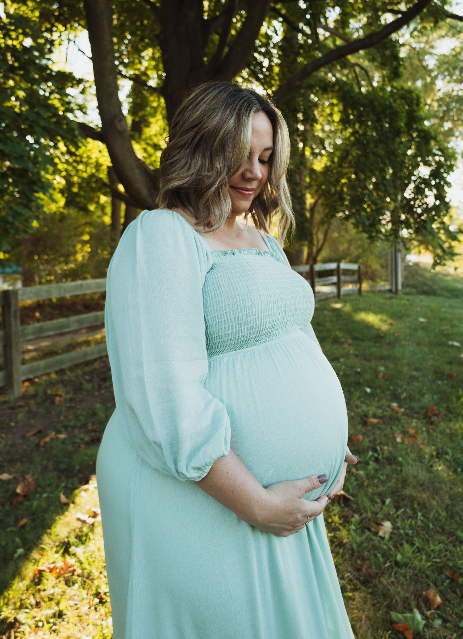 A person in a mint green dress cradles their pregnant belly while standing in a sunlit outdoor setting with trees.