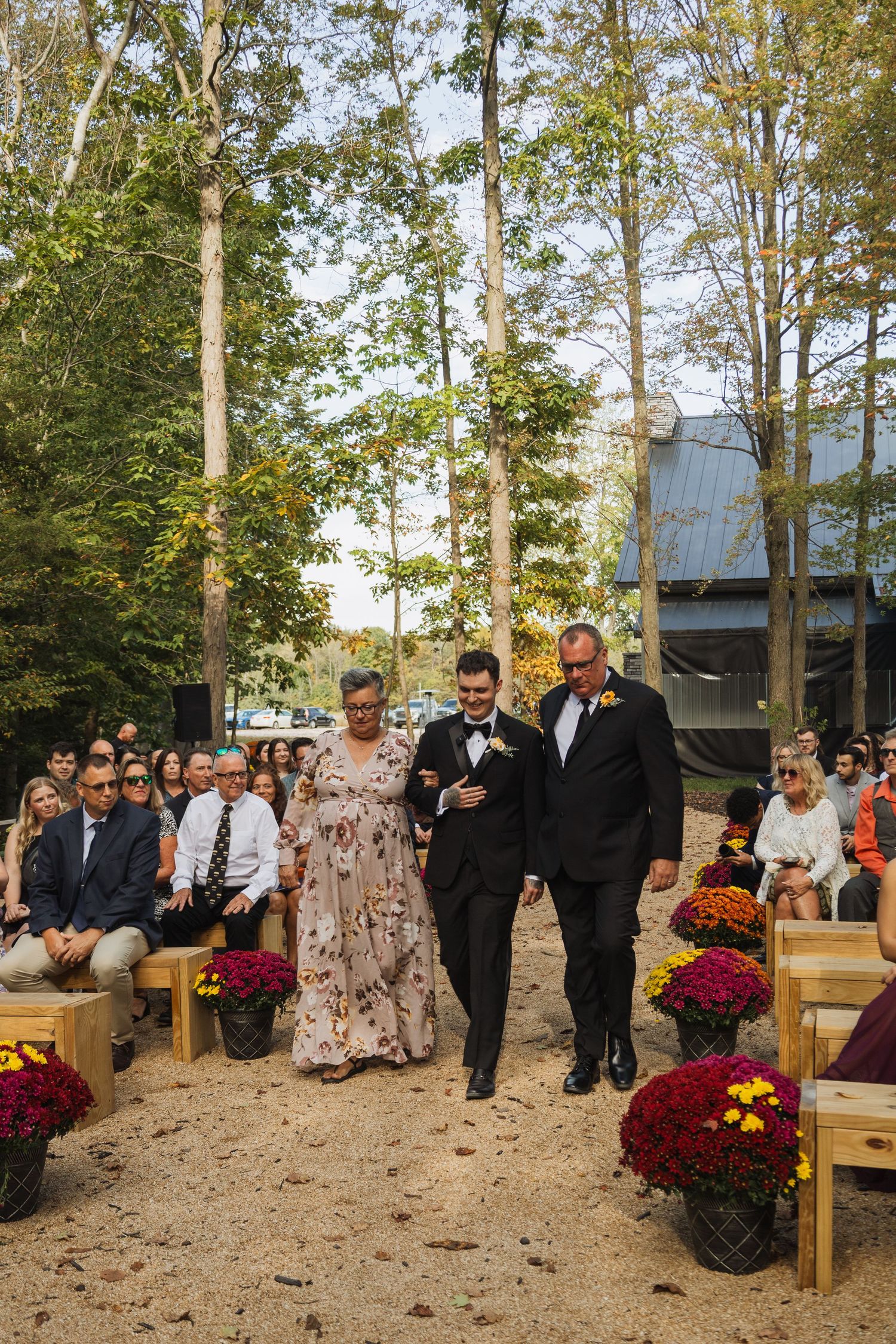Wedding ceremony setup in wooded area with wooden benches and red mum flowers.