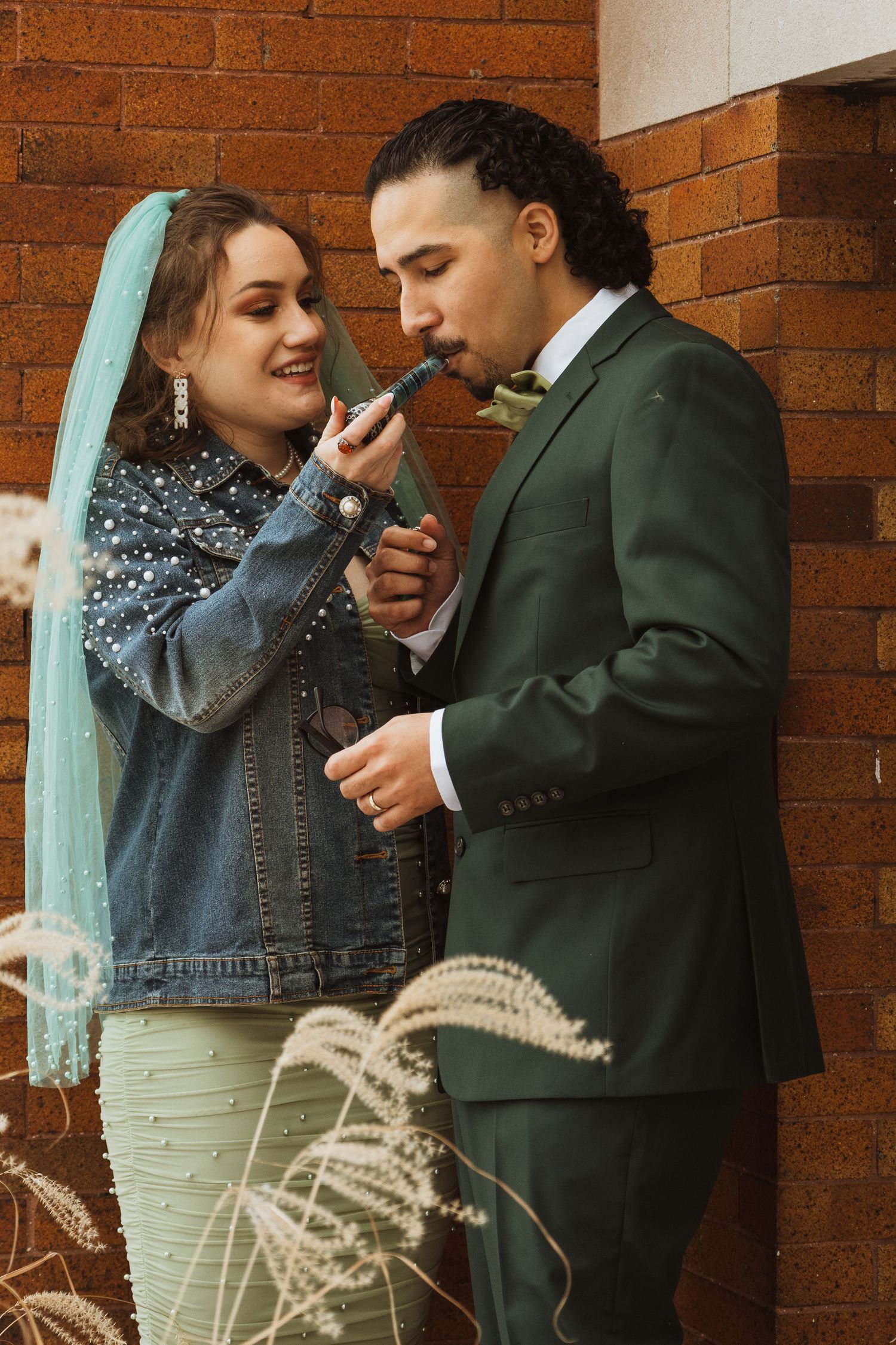 Smiling bride in green veil feeds groom something outside a rustic brick wall.