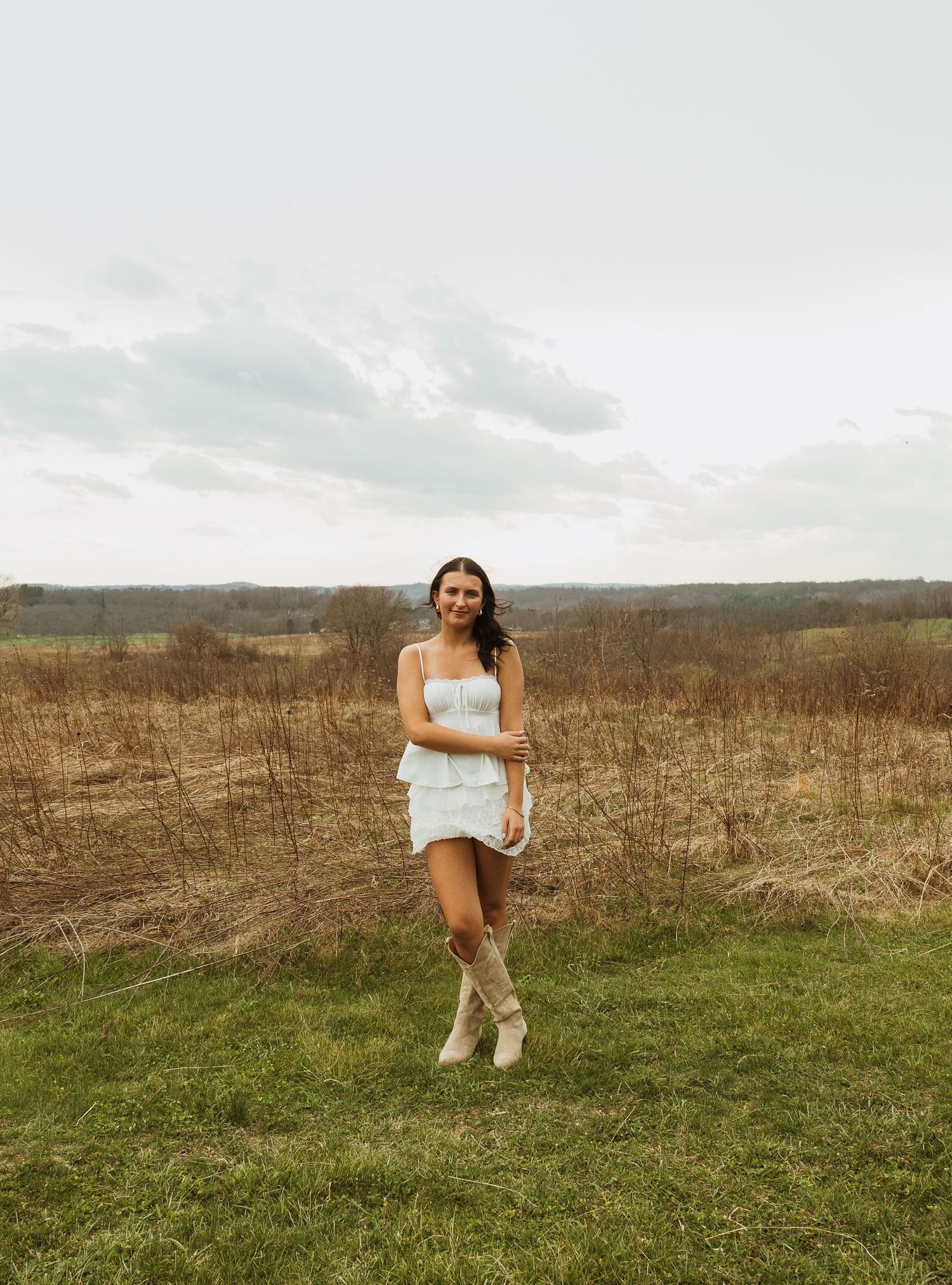 Woman in white mini dress and tan boots posing in a grassy field with overcast sky.