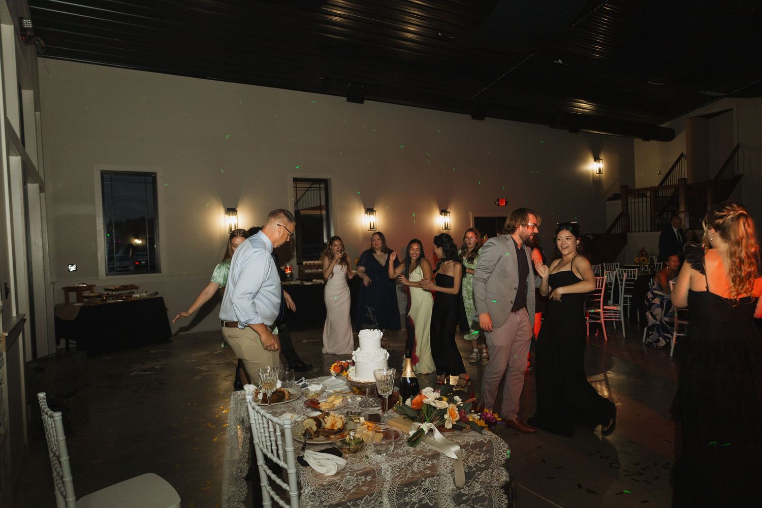 Wedding reception guests dancing and mingling around decorated tables in a dimly lit venue.