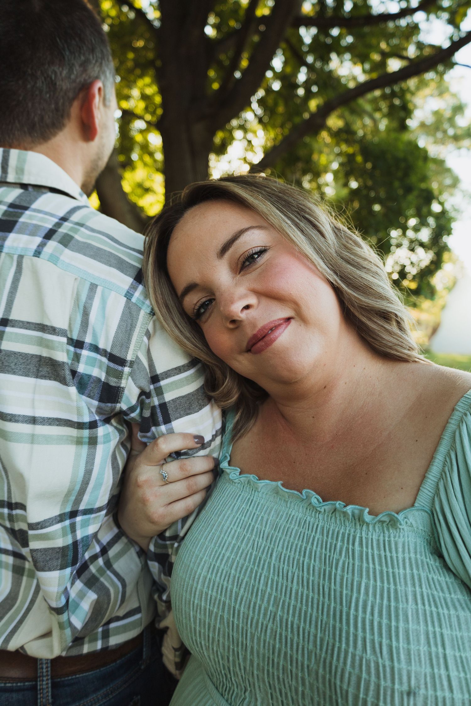 A couple shares a tender moment outdoors in a plaid shirt and mint green top against a sunlit natural backdrop.