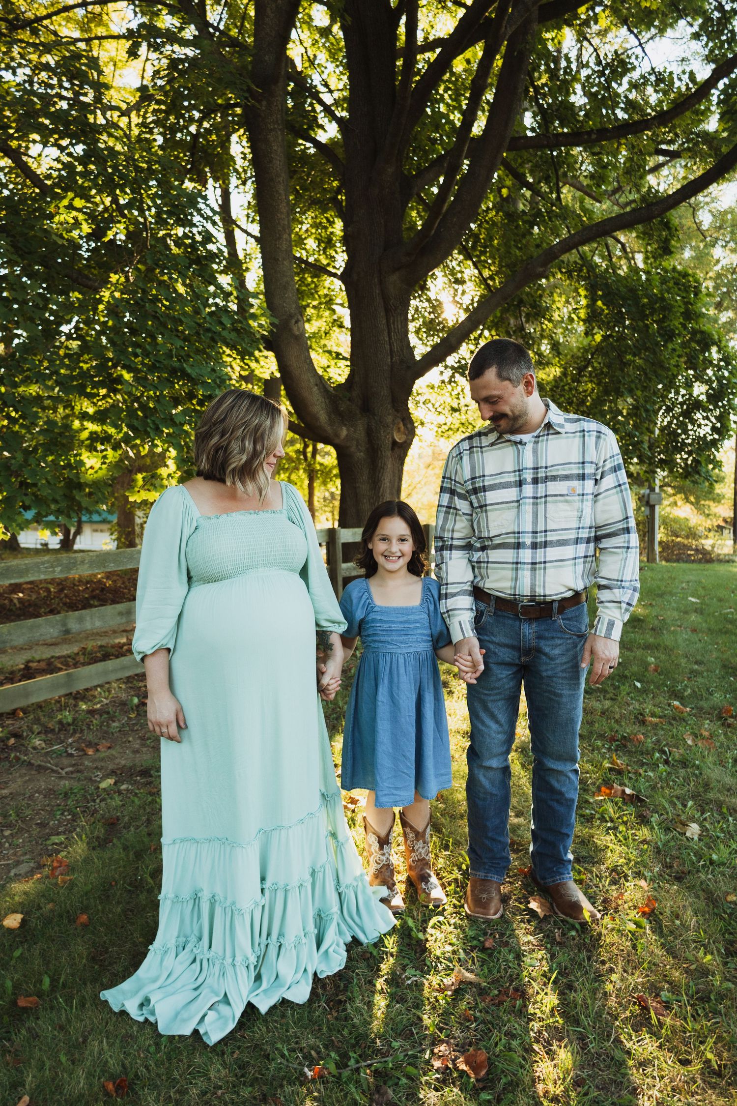 A family stands together in a sunlit garden setting with a wooden fence and large tree in the background.