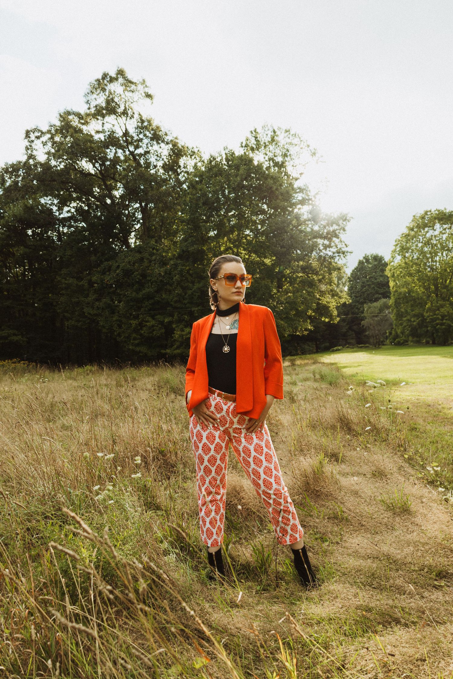 Modern fashion look combining vibrant orange jacket with geometric print pants in outdoor meadow.