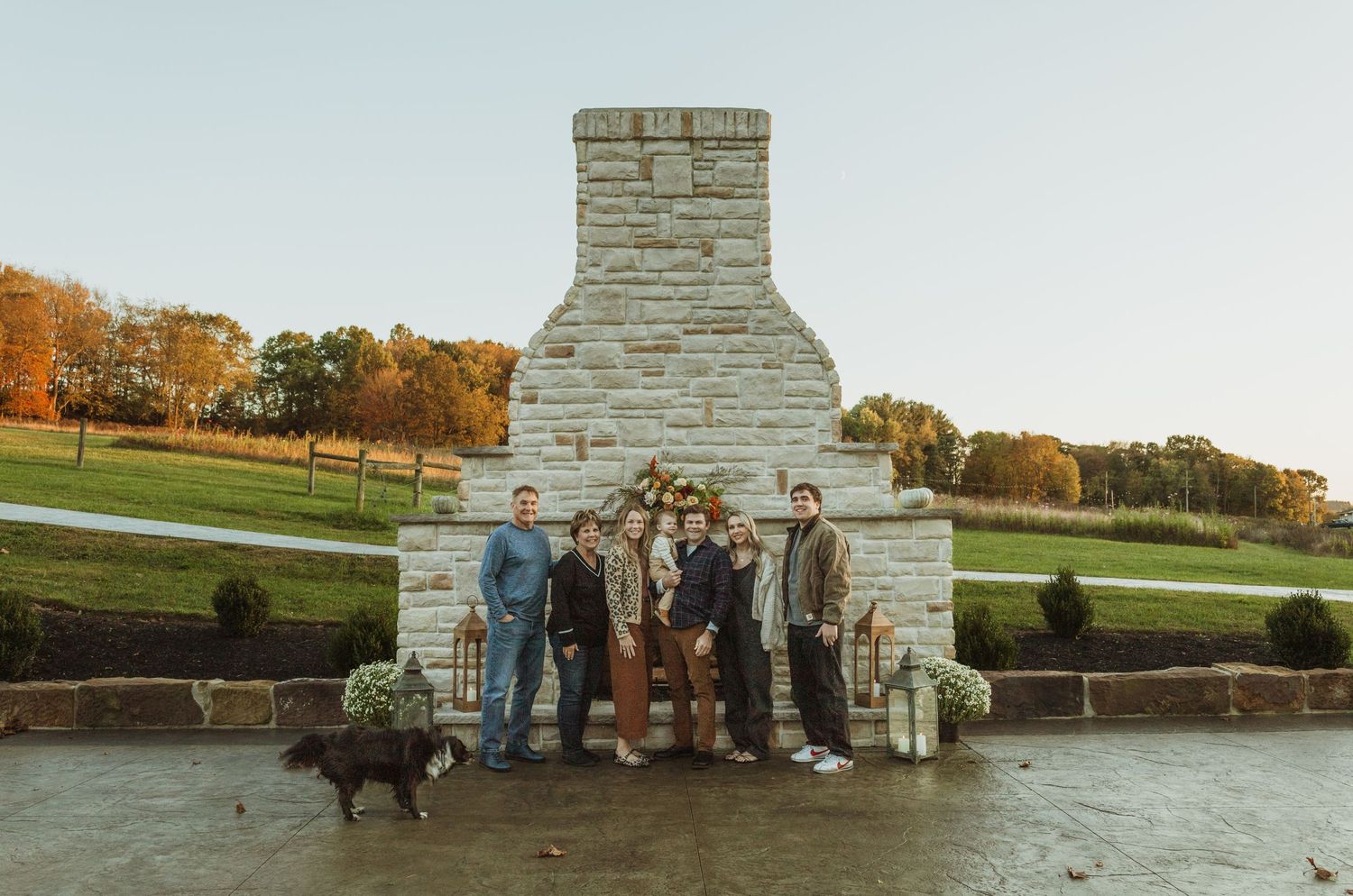 A group gathers in front of a large stone fireplace structure outdoors during autumn.