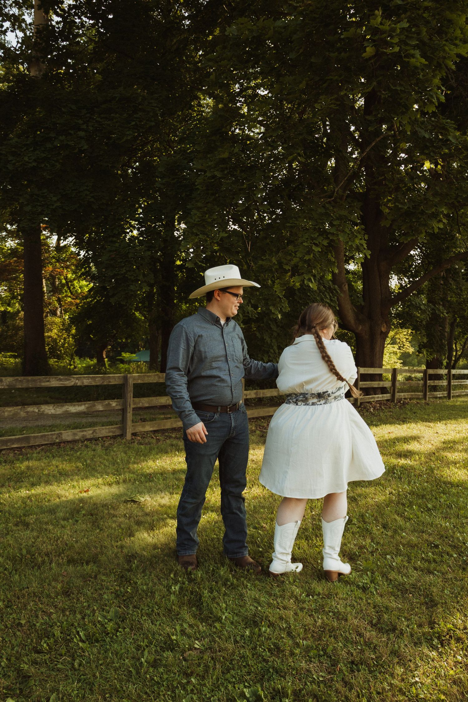 Two people stand together on a grassy hillside surrounded by trees in warm evening light.