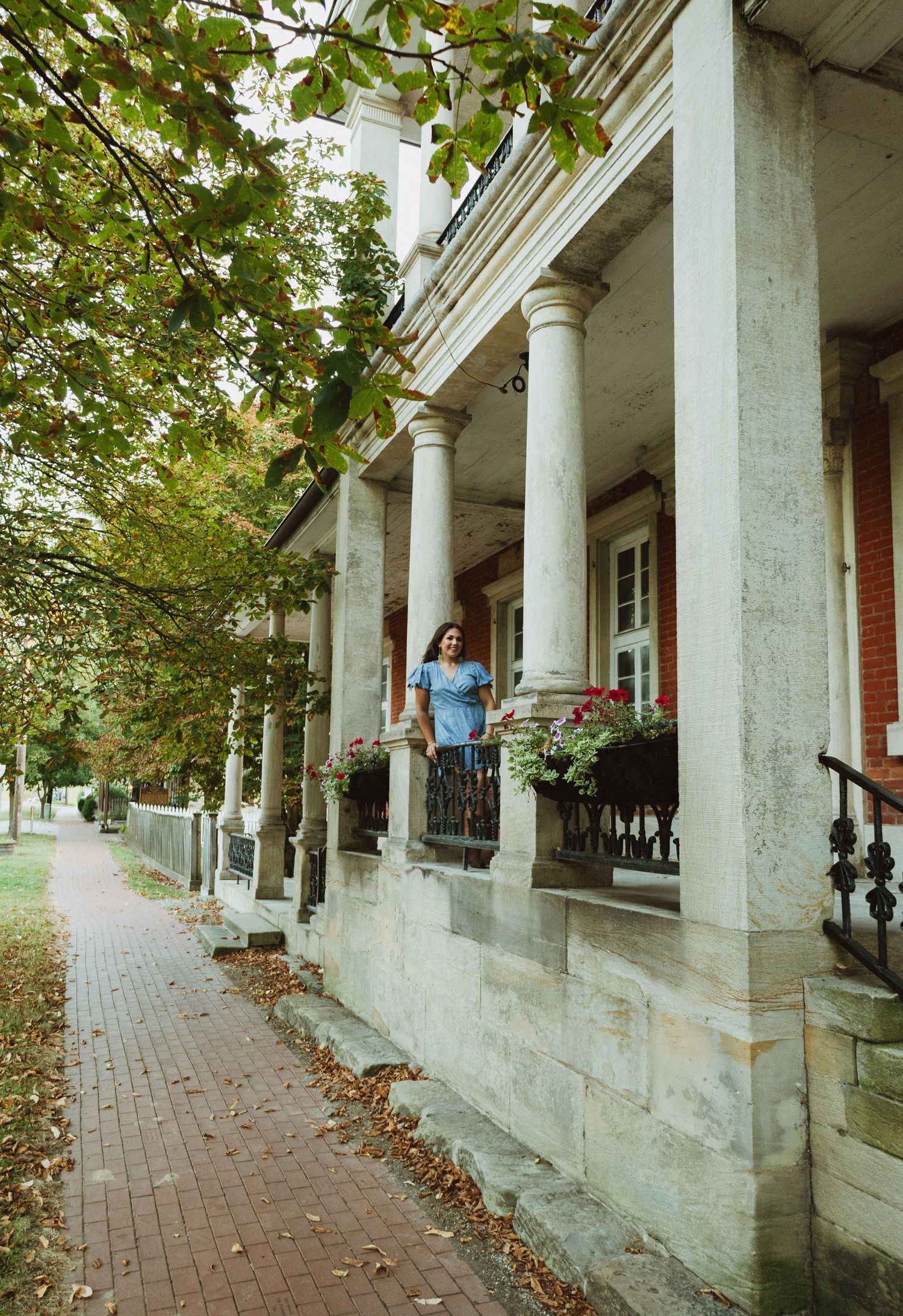 White stone colonnade with tall columns extends along a brick path surrounded by lush green trees.