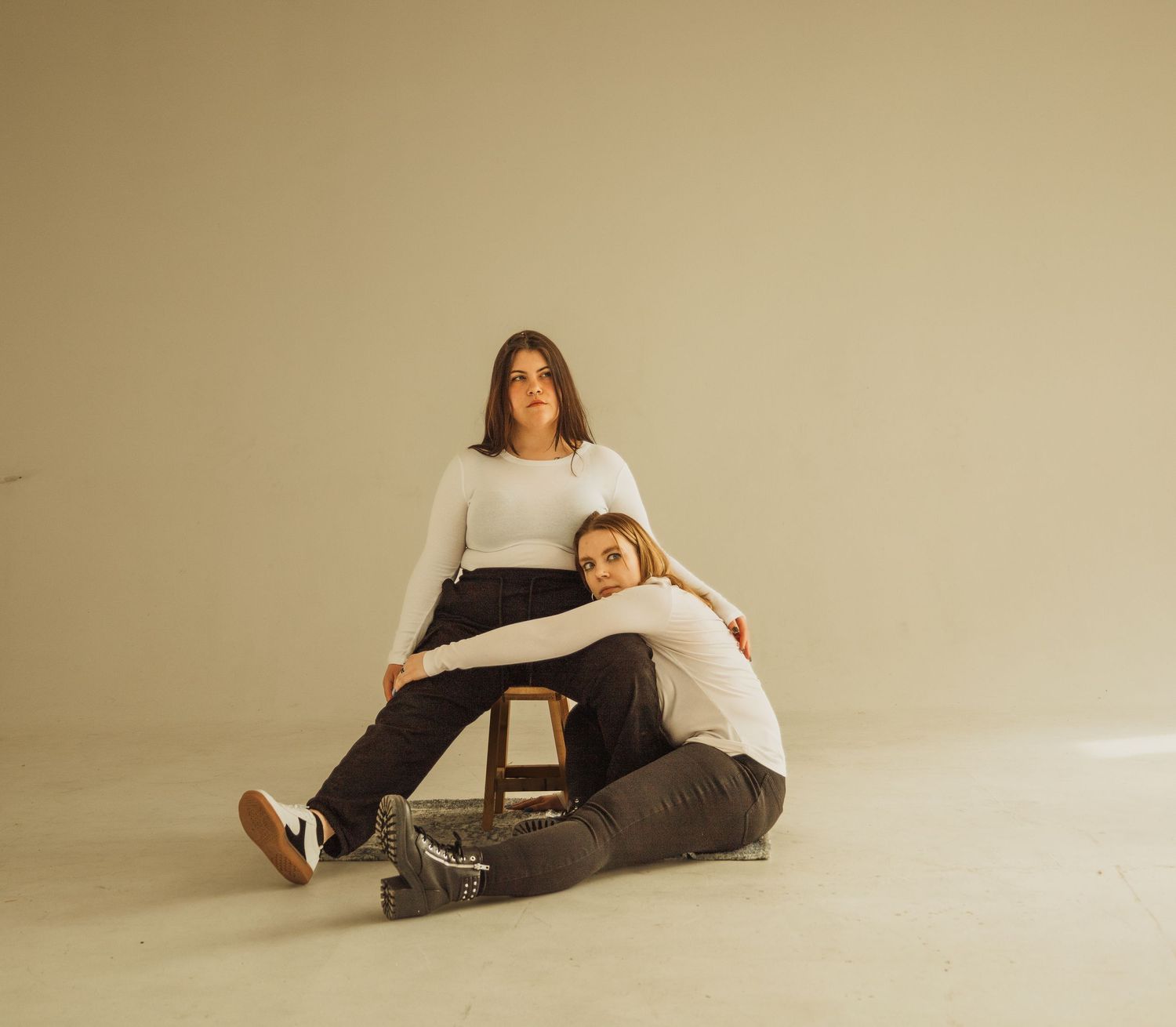 Two people in casual attire posing with wooden chair in minimalist beige studio setting.