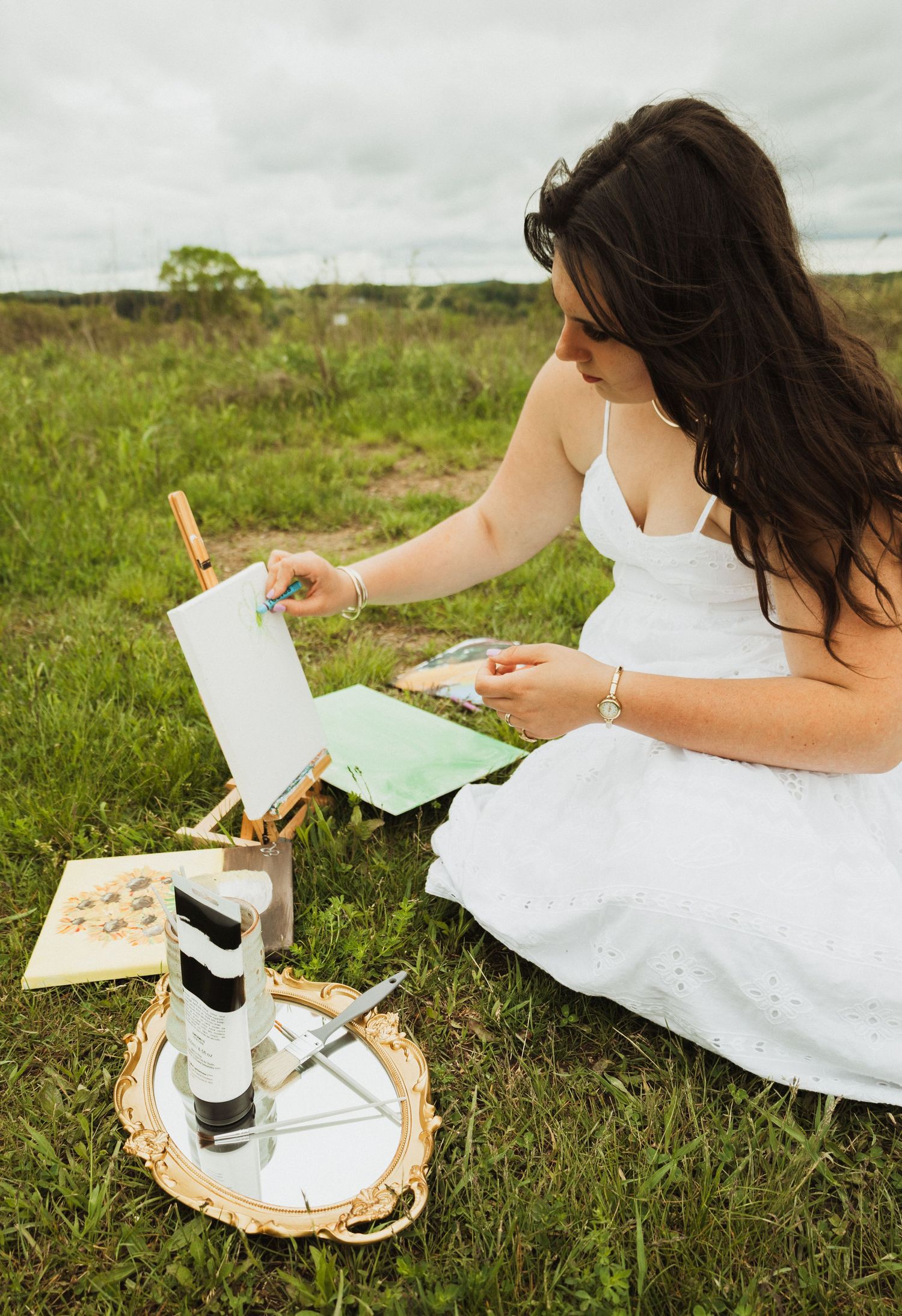 Person in white dress sits in grassy field writing and painting with art supplies and coffee on a woven mat.