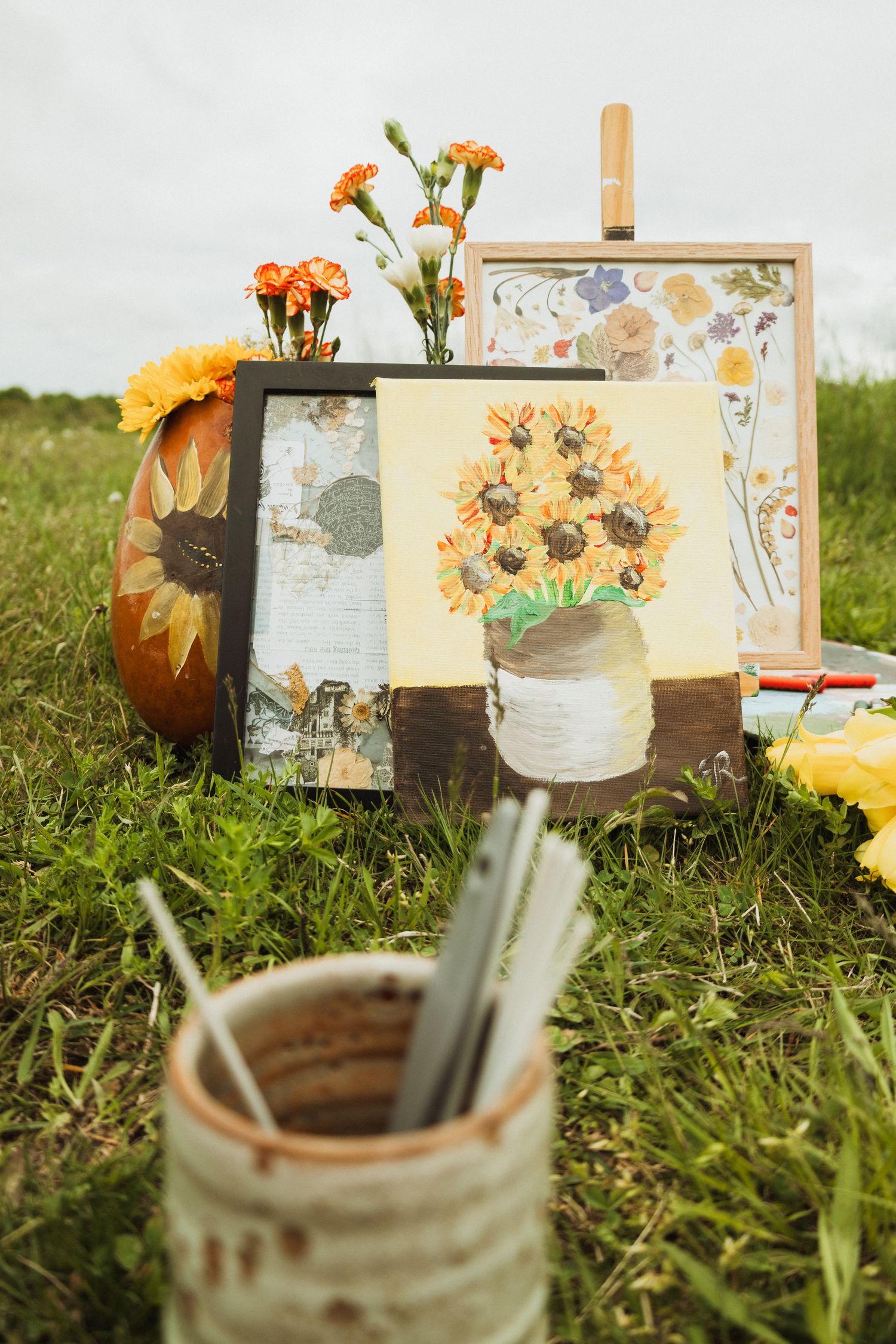 Artistic fall setup with sunflower paintings, orange carnations and art supplies displayed on grass outdoors.