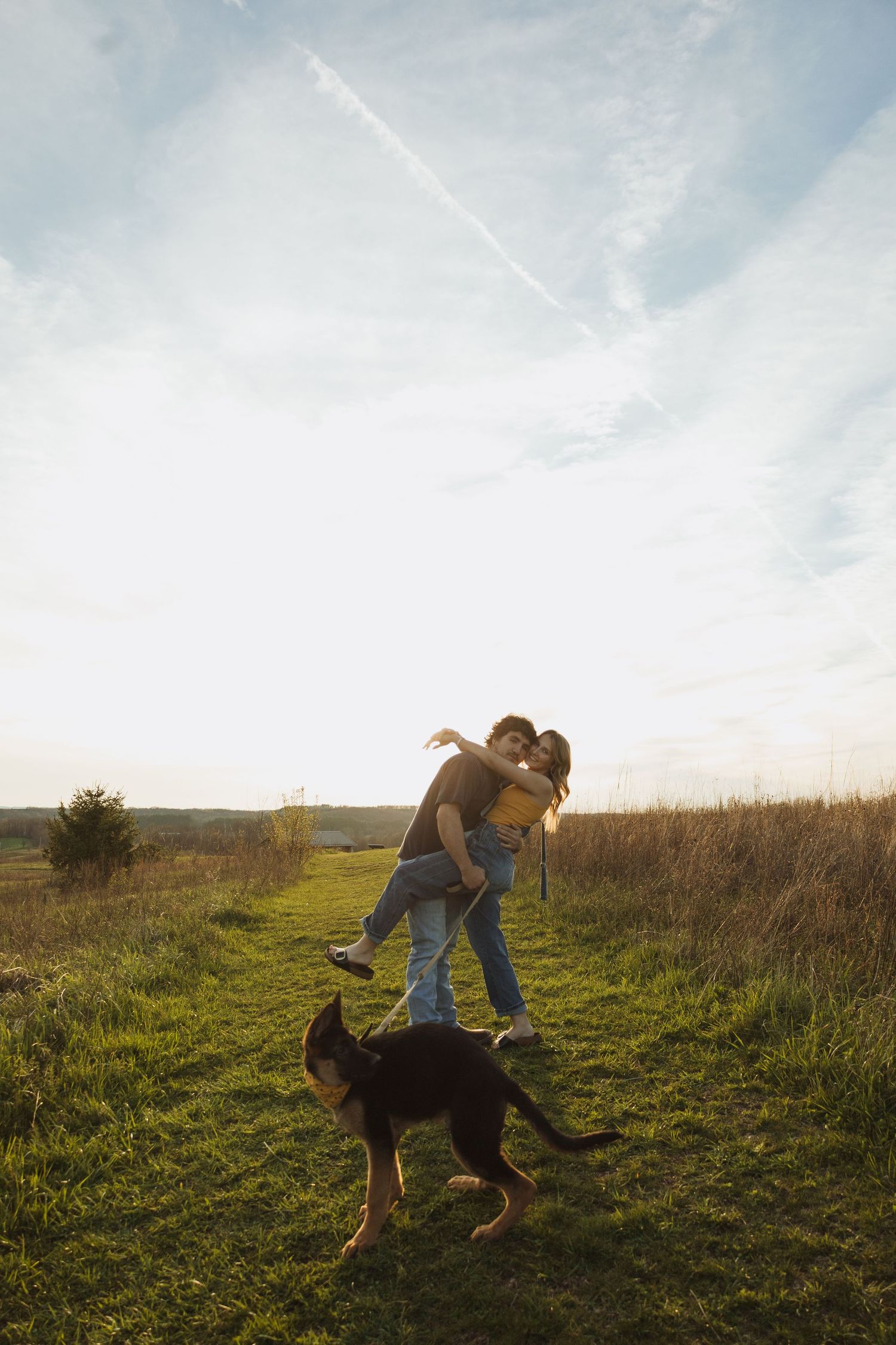 Couple playing with a black dog in a sunny green field, man giving woman a piggyback ride under a blue sky.