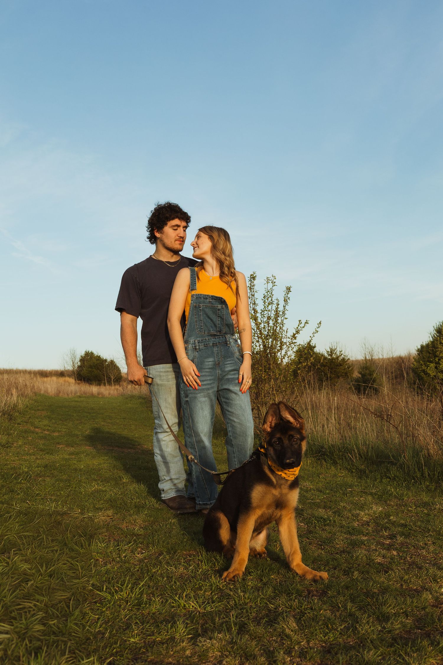Young couple with German Shepherd dog standing in a sunny field at golden hour, woman in denim overalls gazing up.