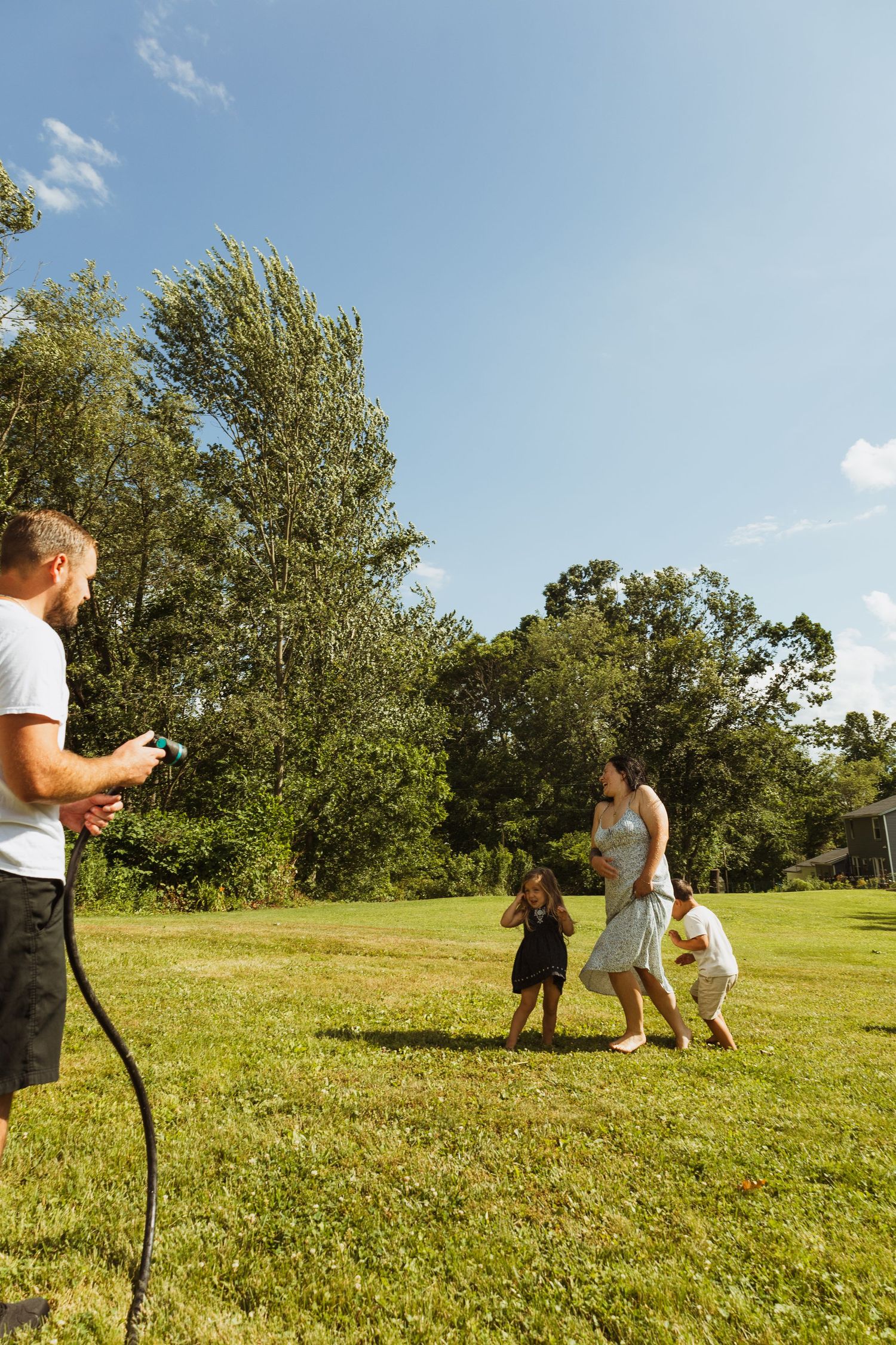Family playing with garden hose on sunny day in green backyard, children running while man sprays water outdoors.