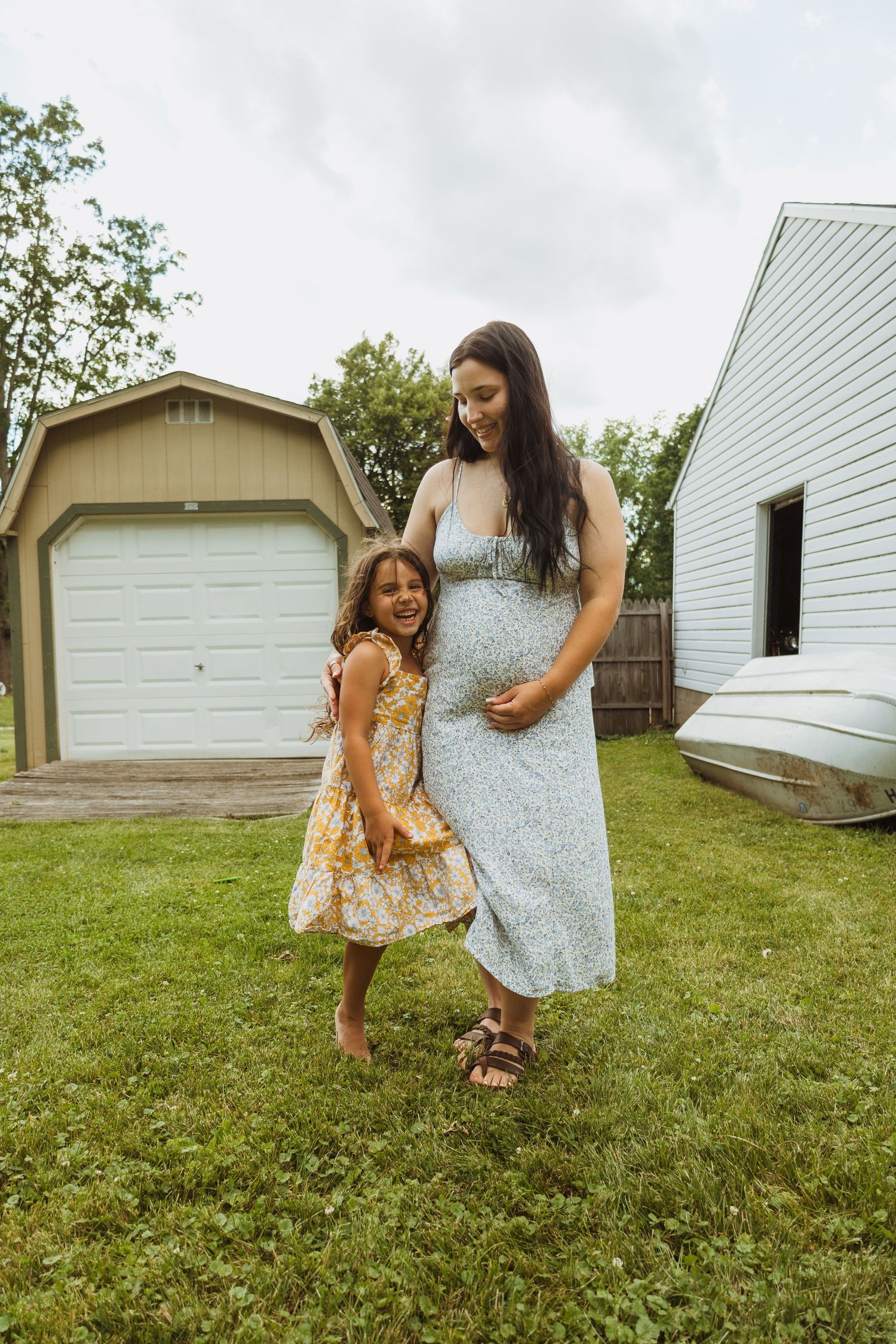 Pregnant mom in blue dress smiles as young girl hugs her baby bump in backyard.