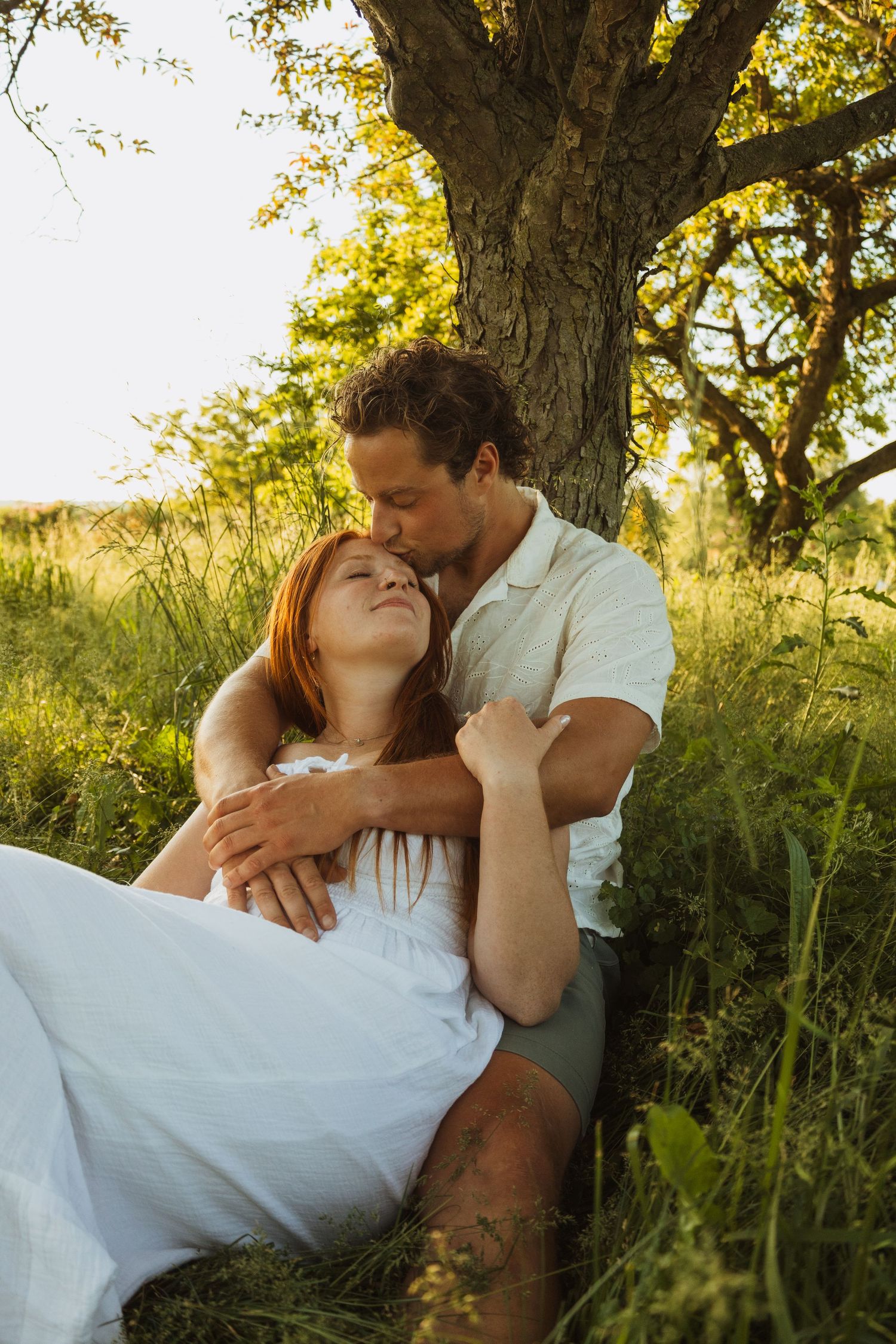 Couple sitting under a tree in golden sunlight, man kissing woman's head while she reclines in his arms.