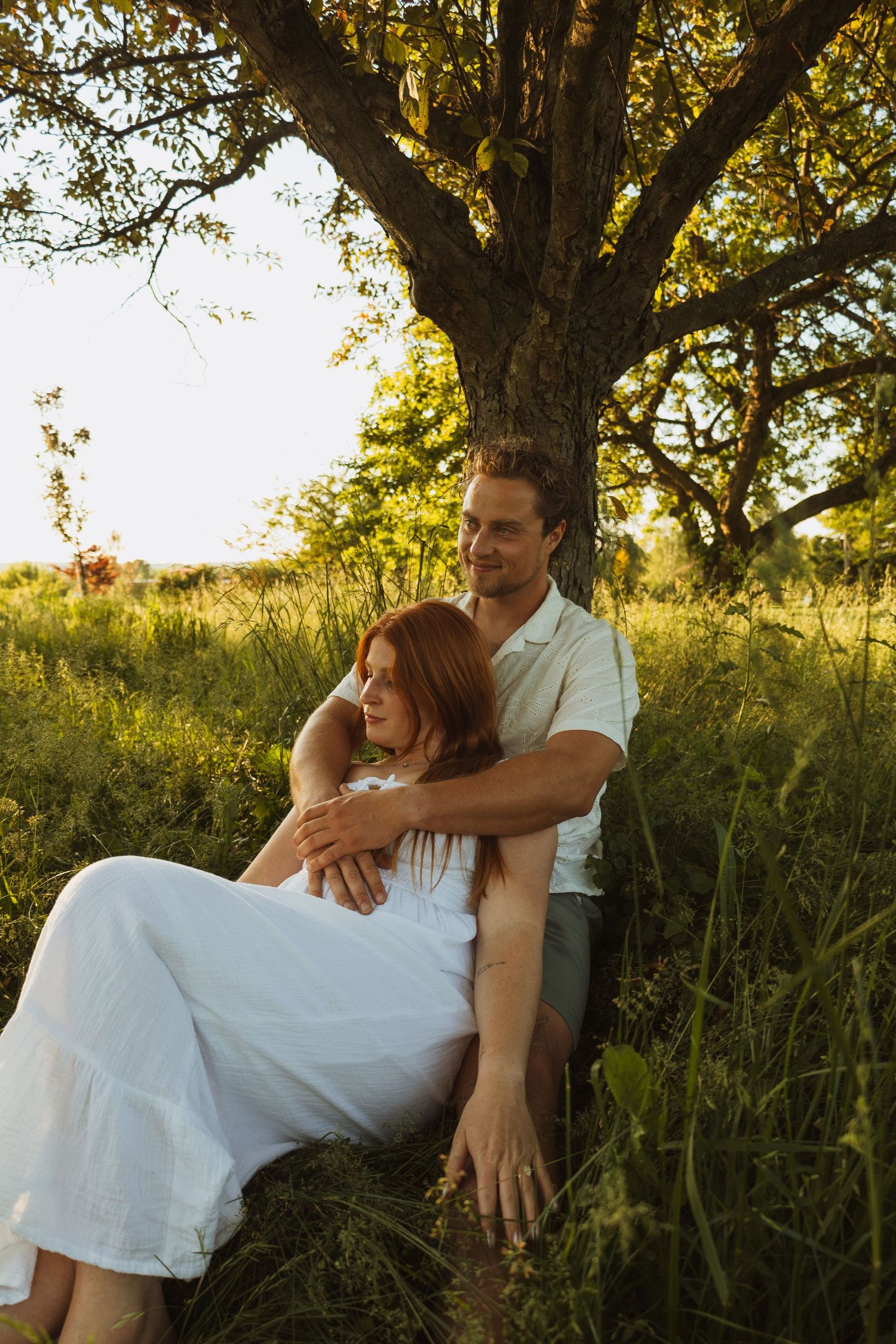 Couple sitting together under a large tree in a sunny green field, woman in white dress leaning against man.