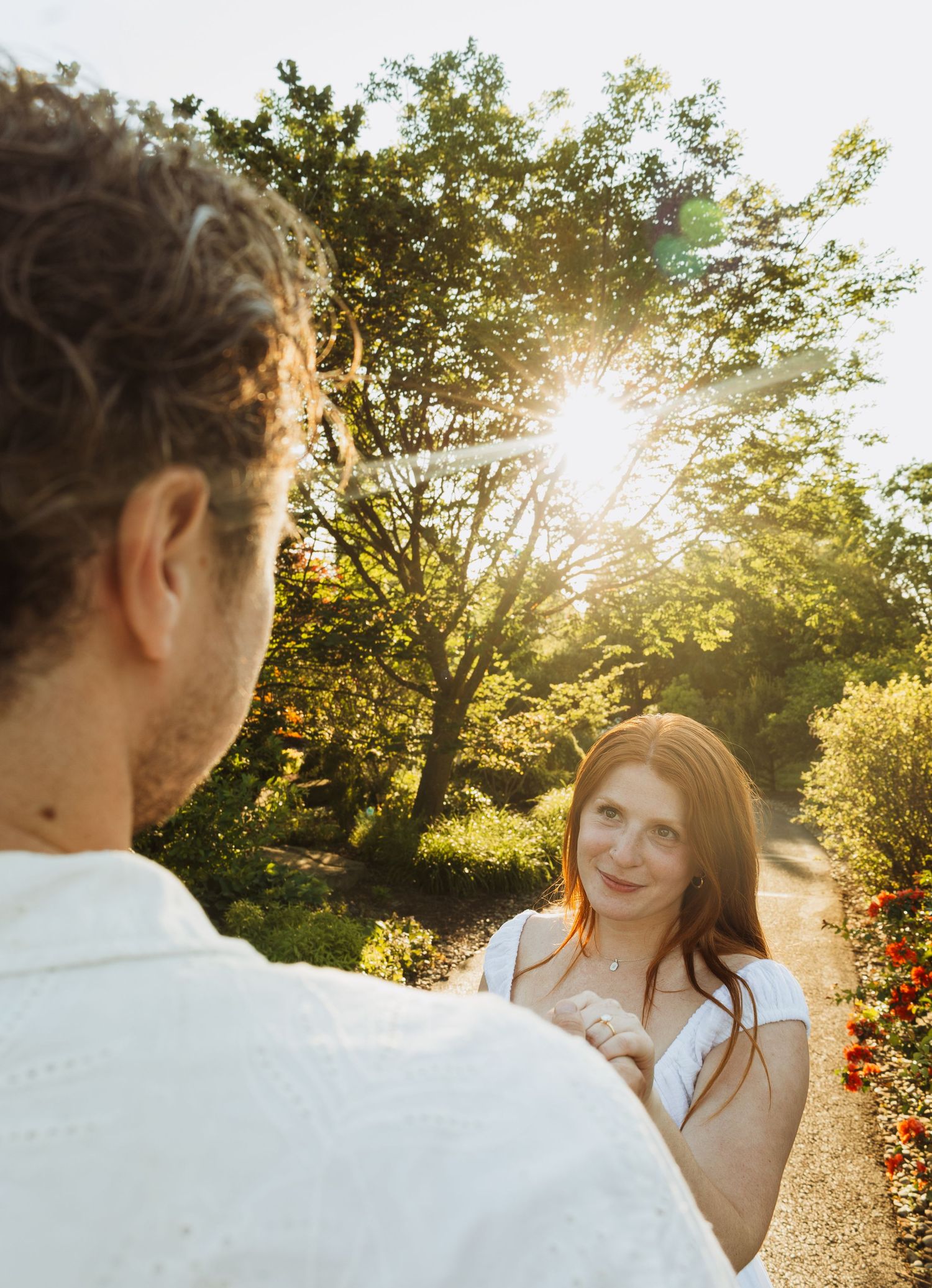 A couple shares a romantic moment in a sunlit garden, with a red-haired woman smiling warmly at a man in a white shirt.