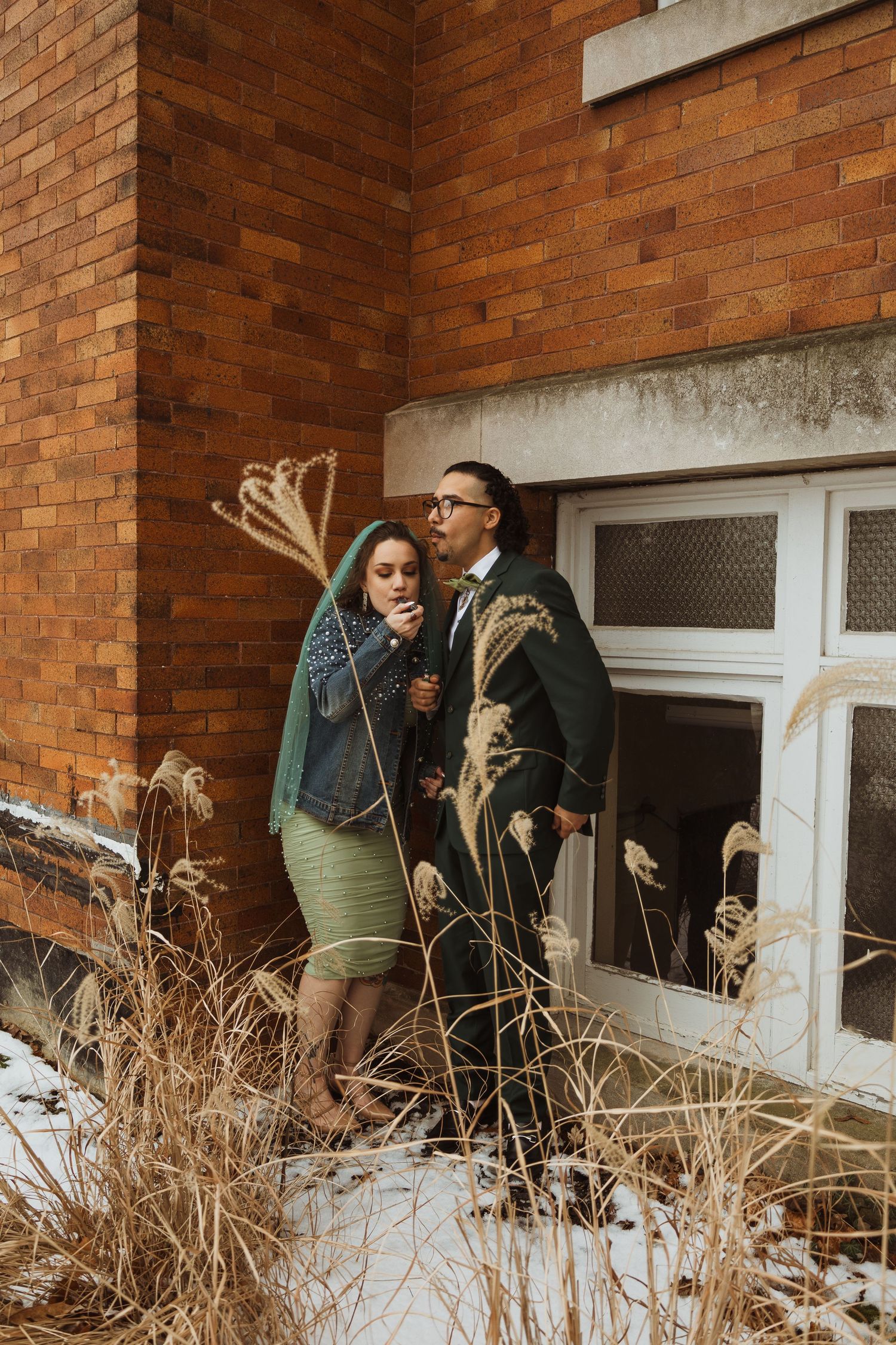 Newlywed couple standing together outside a brick building surrounded by winter grass.