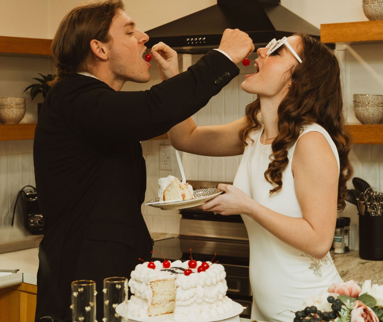 Newlyweds feeding each other wedding cake in a modern kitchen setting.