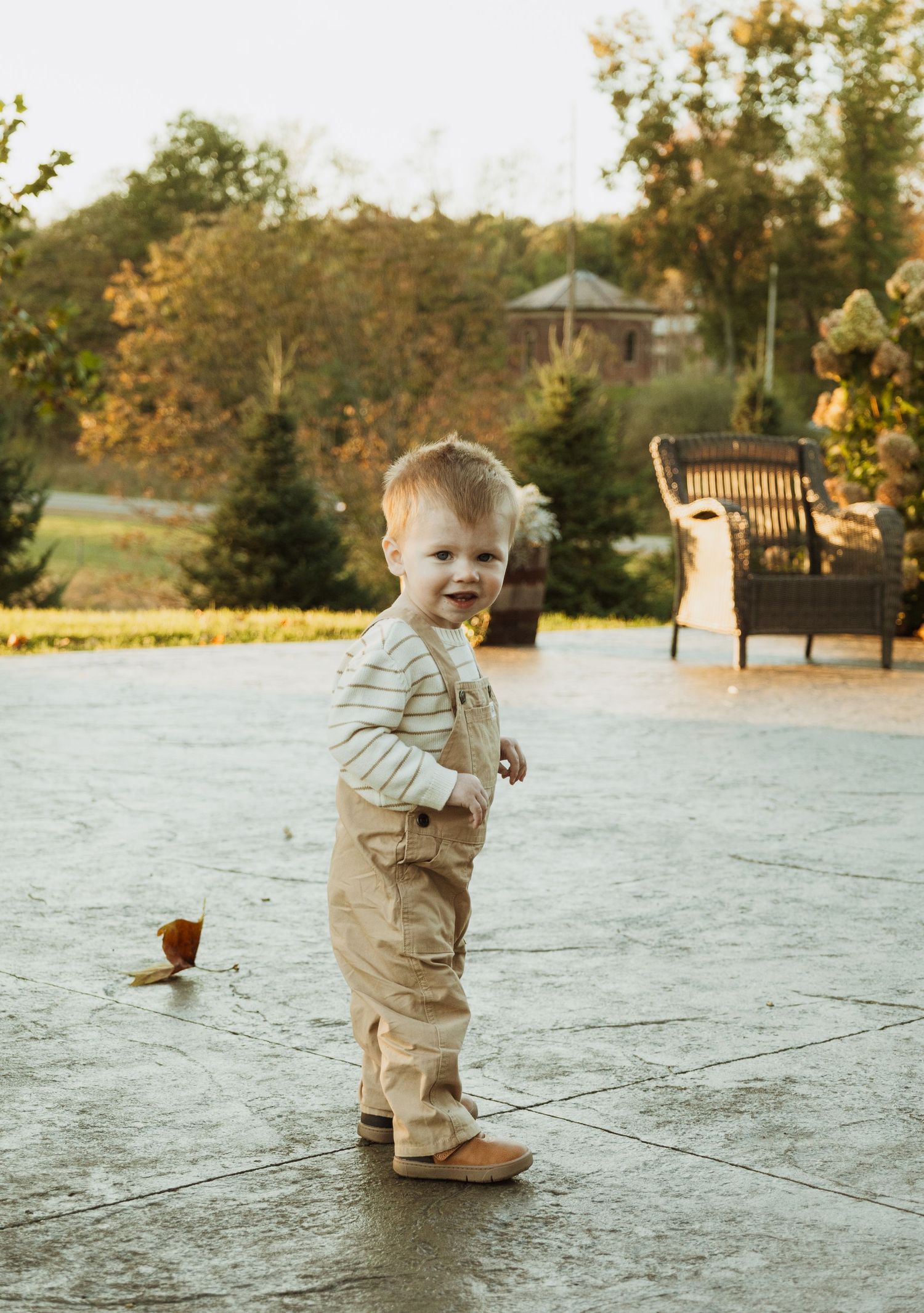 A toddler in khaki pants and grey sweater stands on a stone patio during golden hour at sunset.