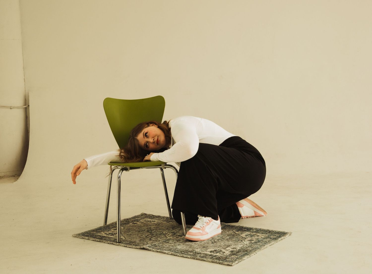 Person resting draped over green chair on patterned rug wearing white shirt and black pants.