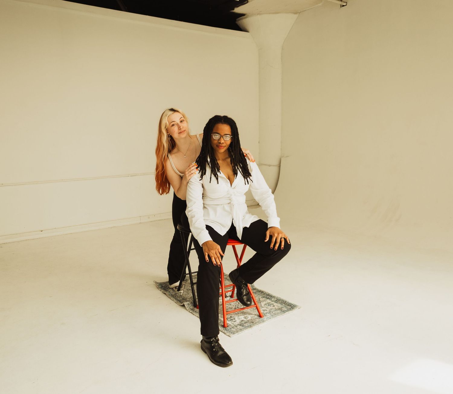 Two friends in black and white outfits pose together in a minimalist white studio setting.