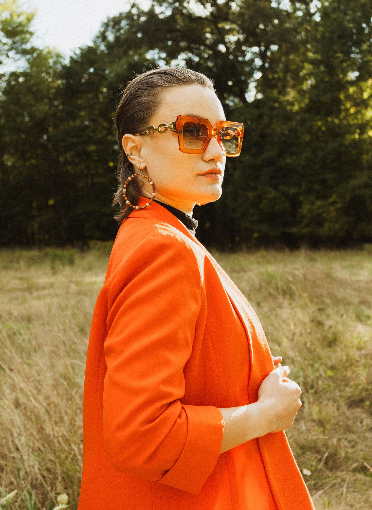 Fashion model in bright orange blazer and retro sunglasses poses in outdoor field during golden hour.